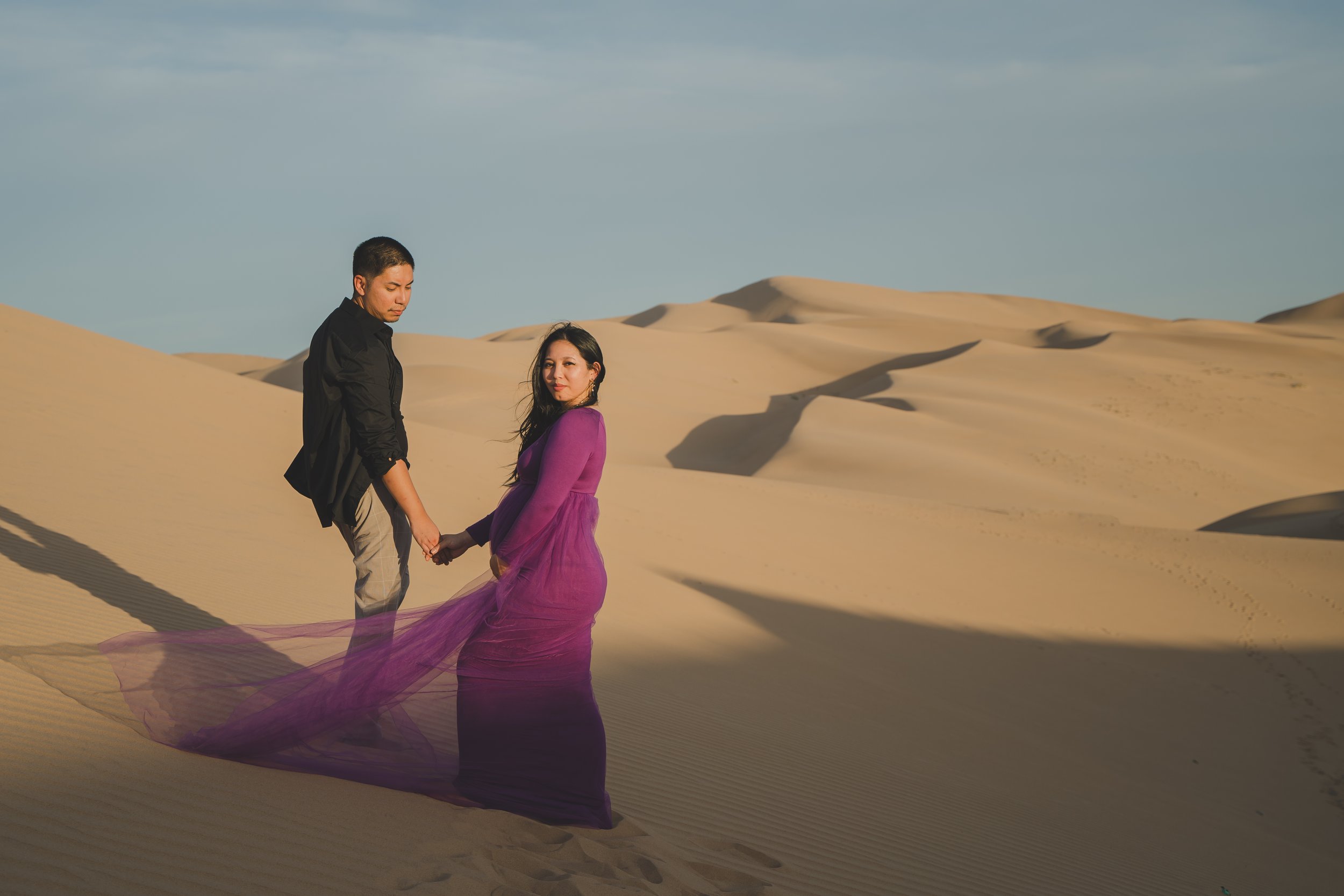 A couple holding hands in a desert landscape with sand dunes and a blue sky, the woman in a flowing purple gown and the man in black clothing.