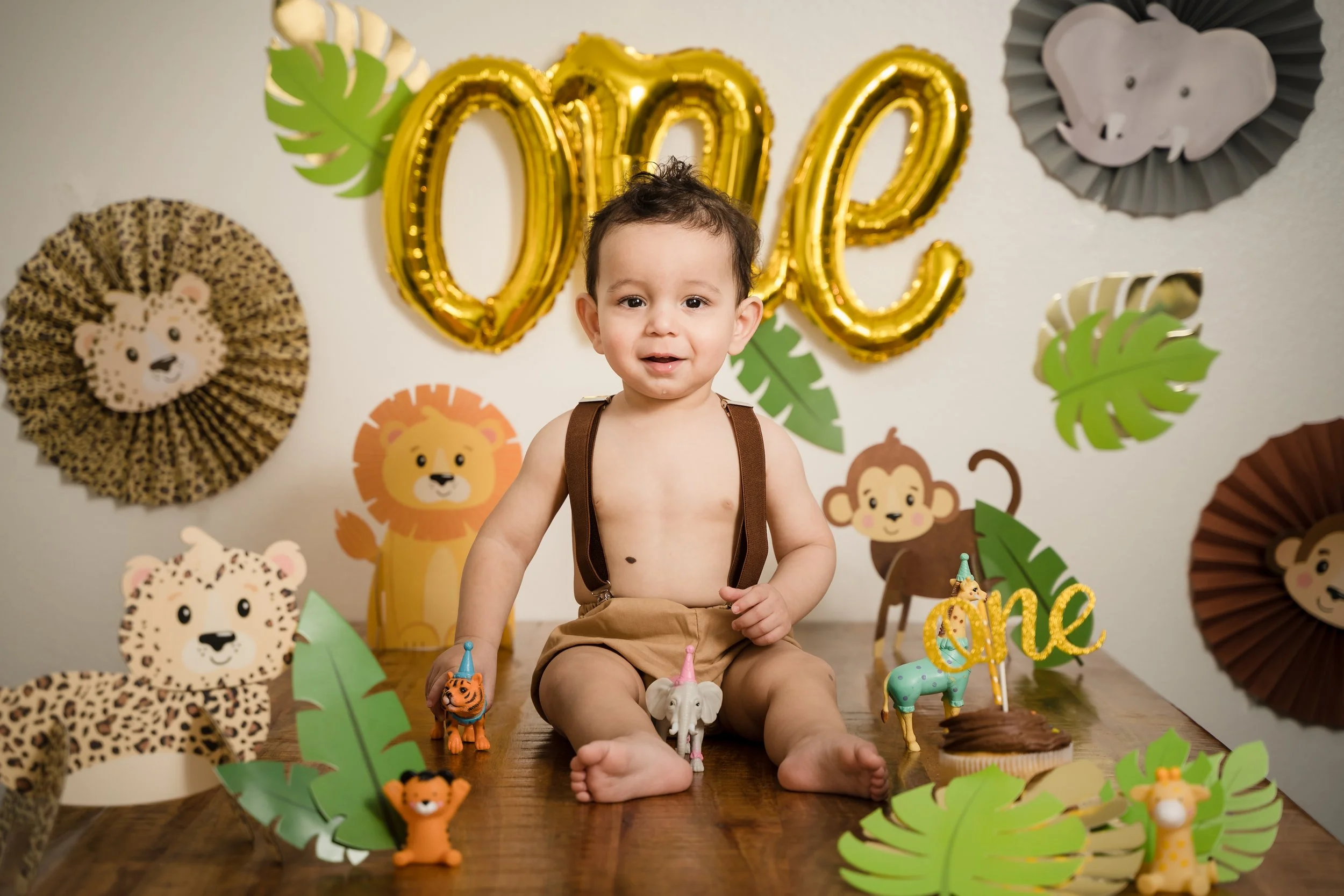Young boy sitting on the floor during a jungle-themed birthday party, with gold balloons spelling '200' and jungle animal decorations including lions, leopards, tigers, and elephants, along with green paper leaves.