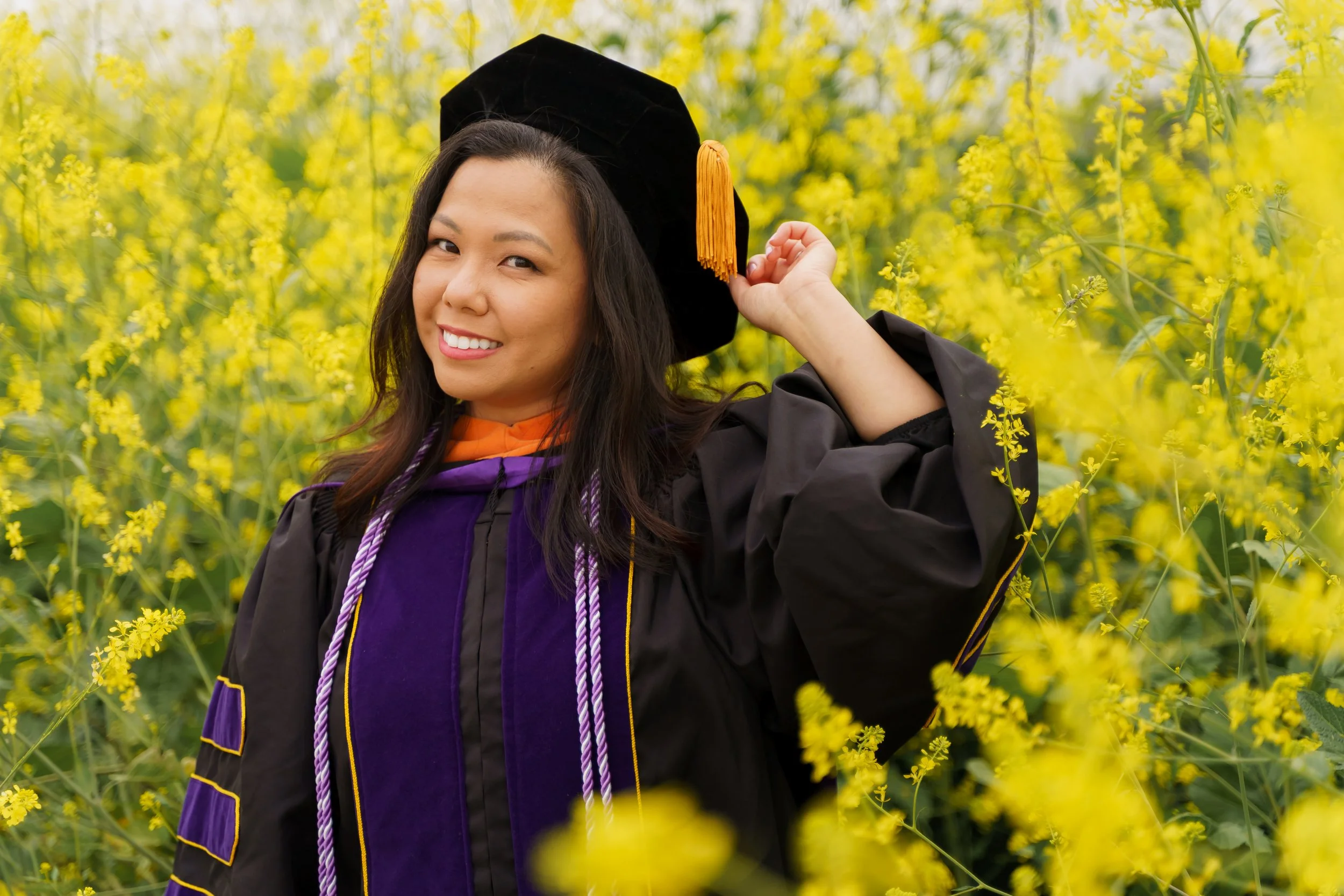 A woman in graduation robes and cap with orange tassel standing among yellow flowering plants, smiling at the camera.
