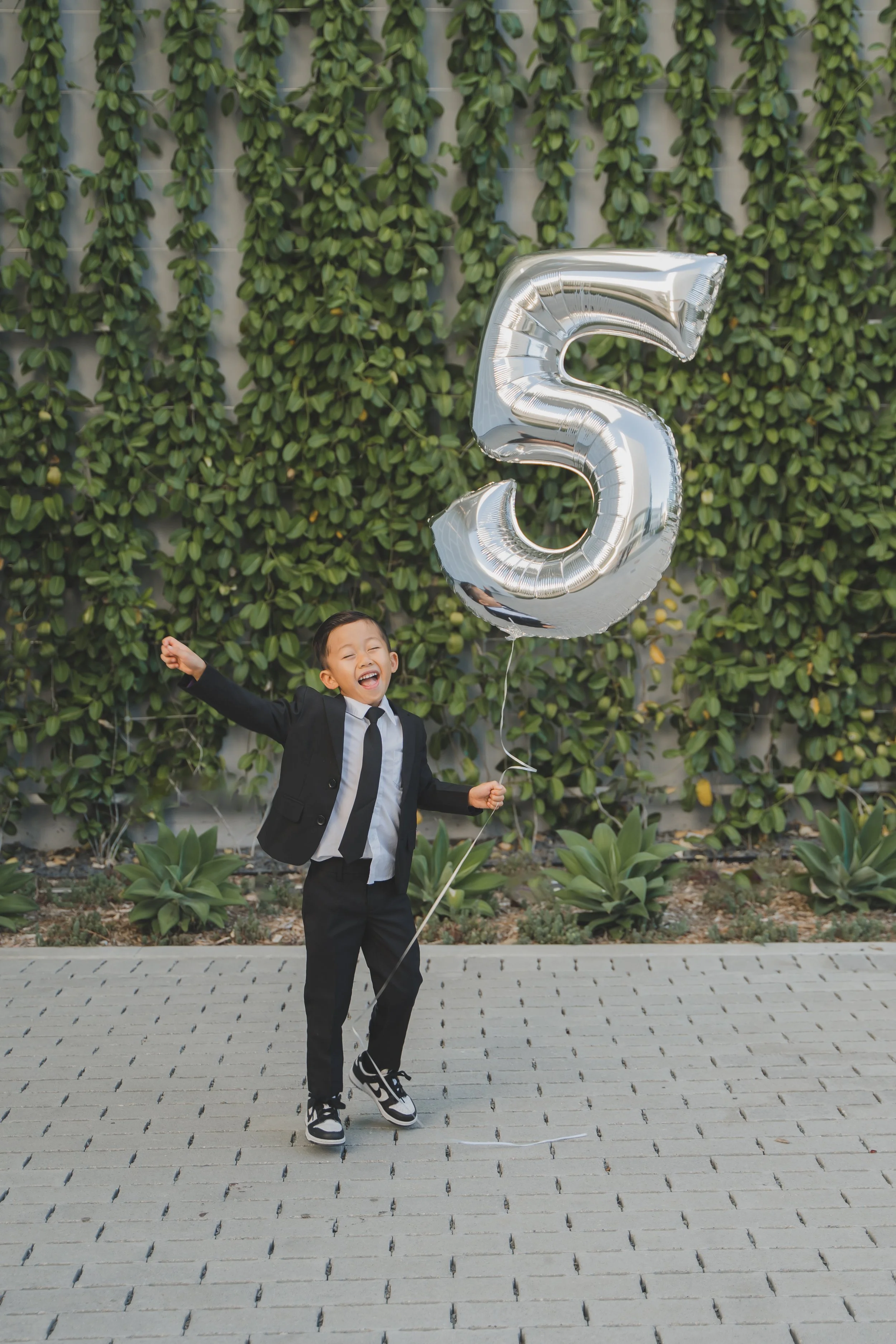 A young boy in a black suit holding a large silver helium balloon shaped like the number 5. He is smiling and appears to be celebrating.