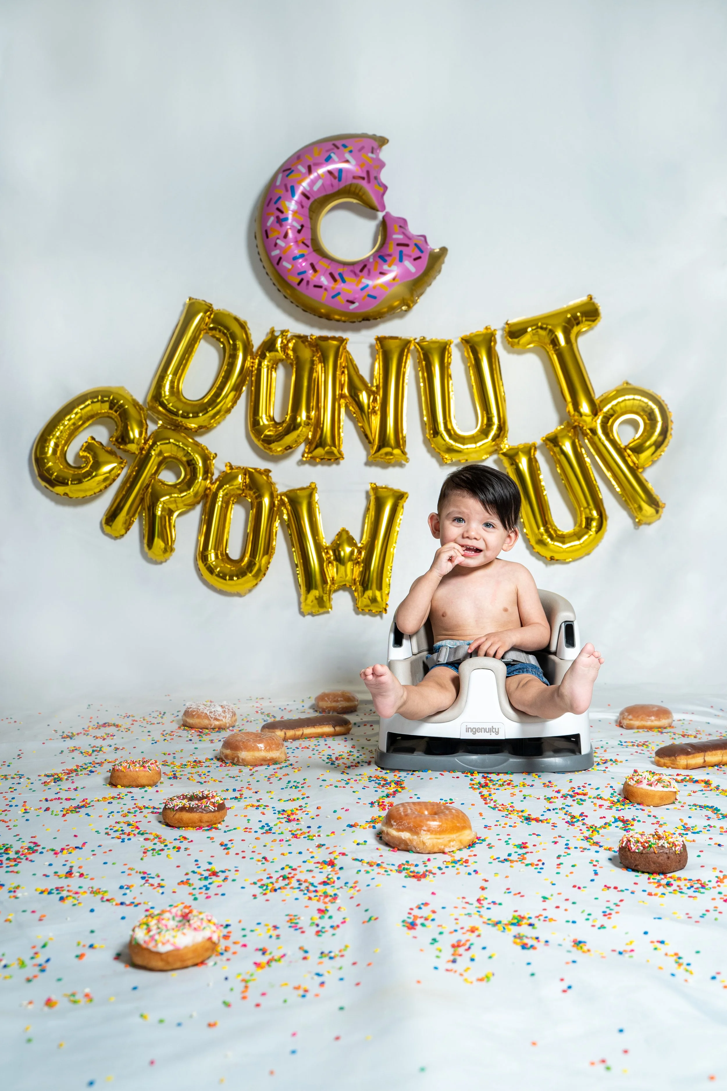 A young boy sitting in a high chair surrounded by scattered donuts and colorful sprinkles, with gold balloon letters spelling 'Sunday Funday' and a pink donut-shaped balloon with sprinkles in the background.