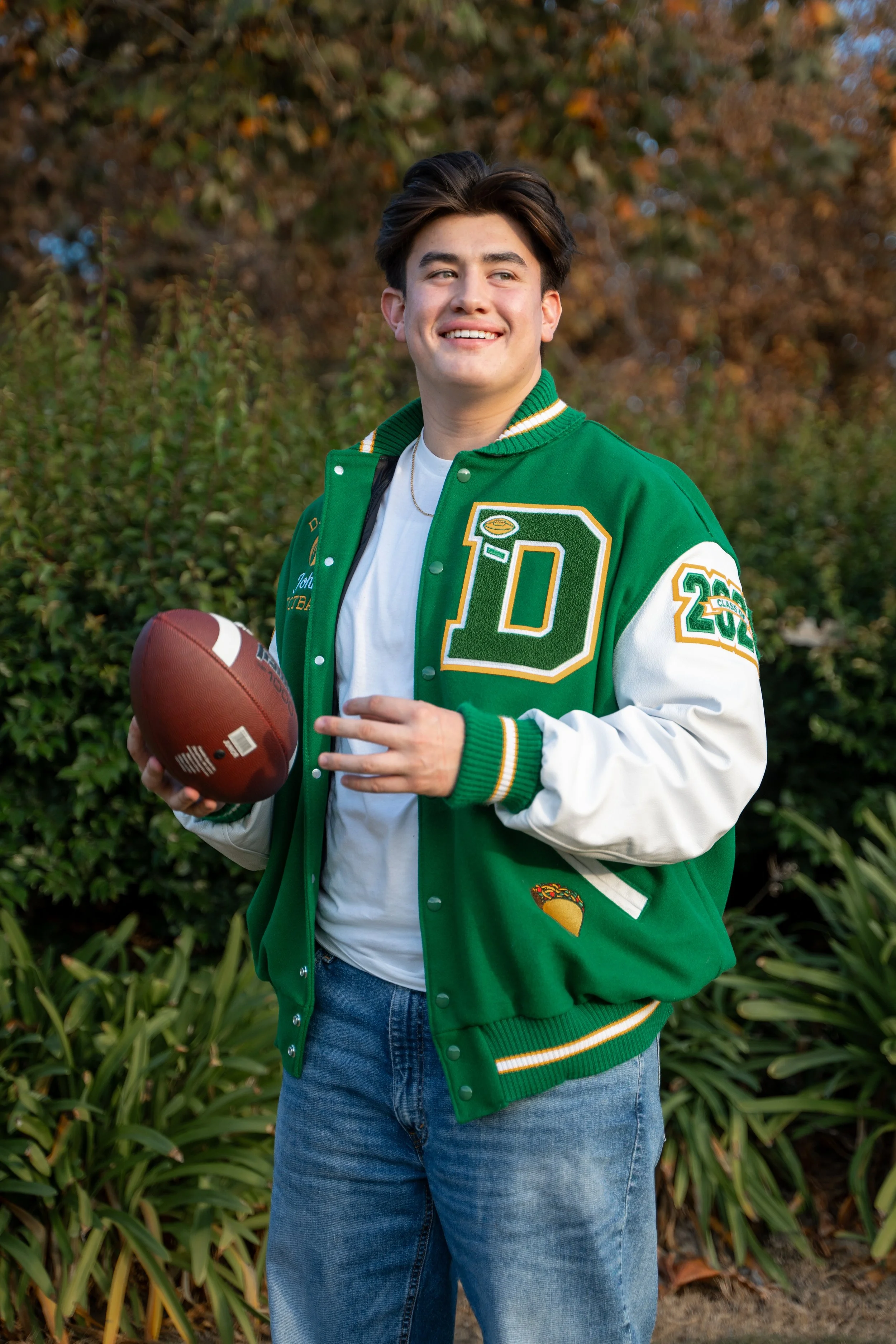Young man smiling outdoors, holding a football, wearing a green and white varsity jacket with patches, jeans, and a white t-shirt, surrounded by green foliage and autumn-colored trees.