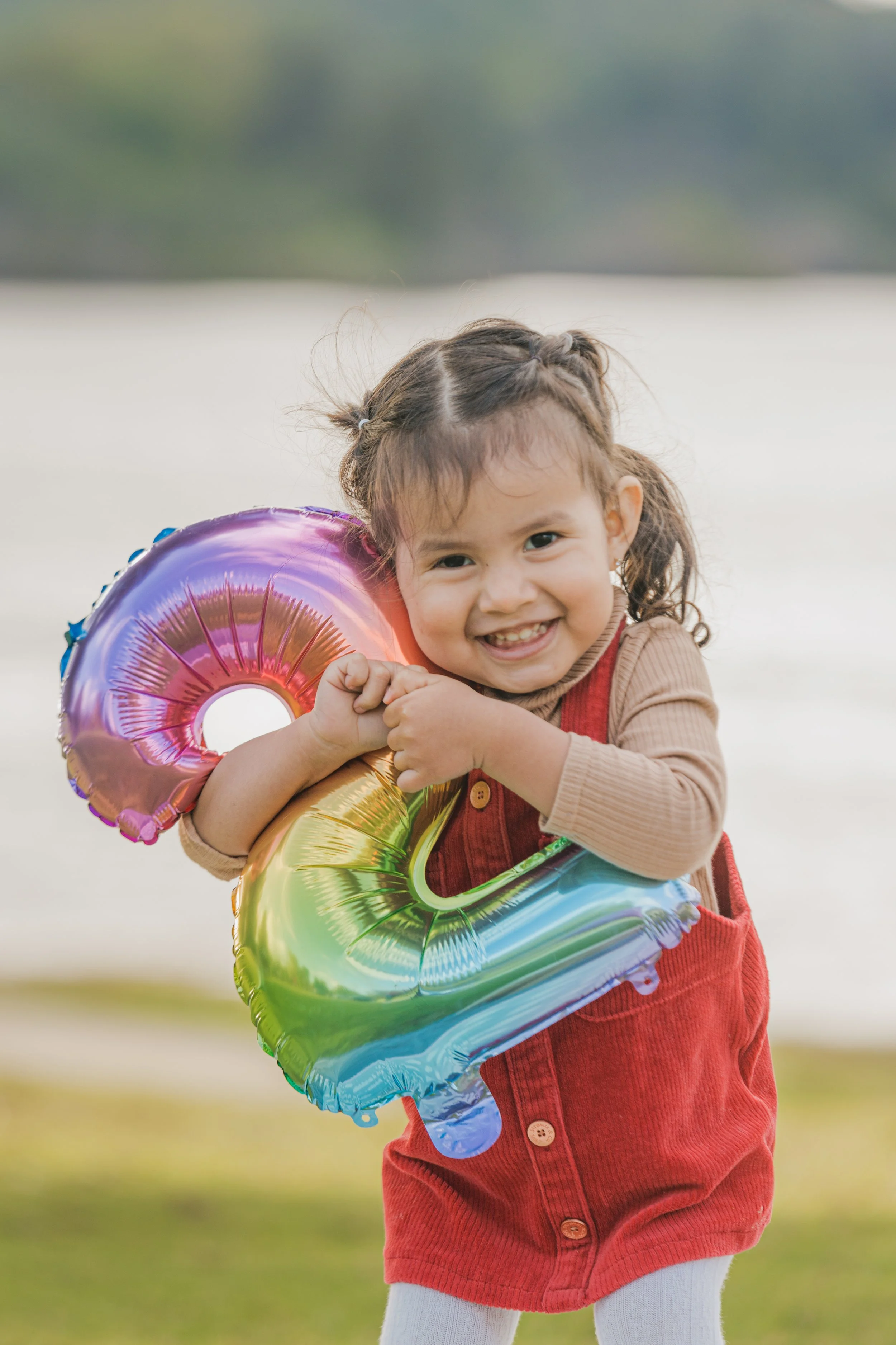 A young girl with curly hair and a big smile, holding a colorful metallic rainbow-shaped balloon, standing outdoors with a blurred natural background.