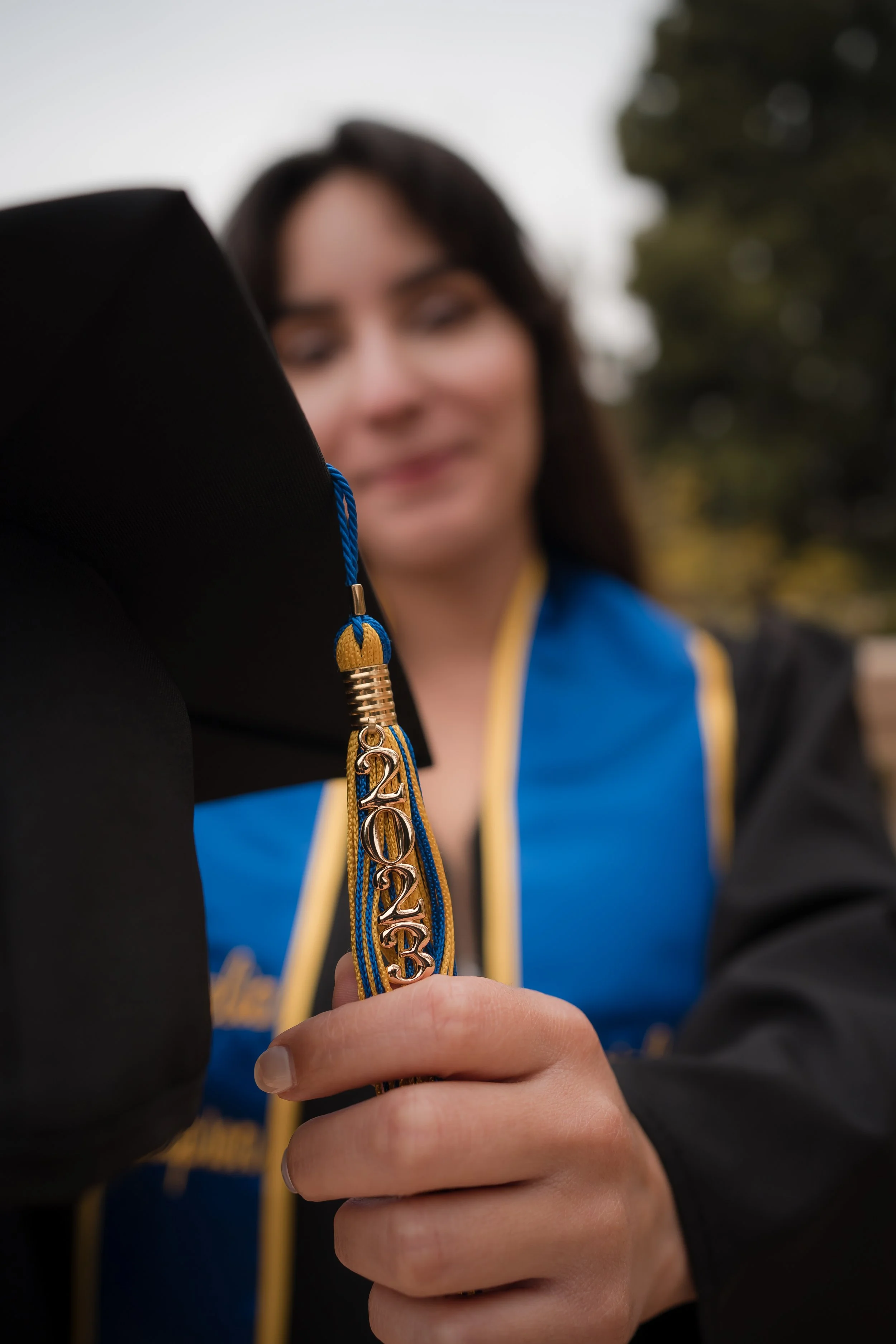 Close-up of a person's hand holding a graduation cap tassel with year '2023' in gold numbers, blurred person in graduation gown in background.