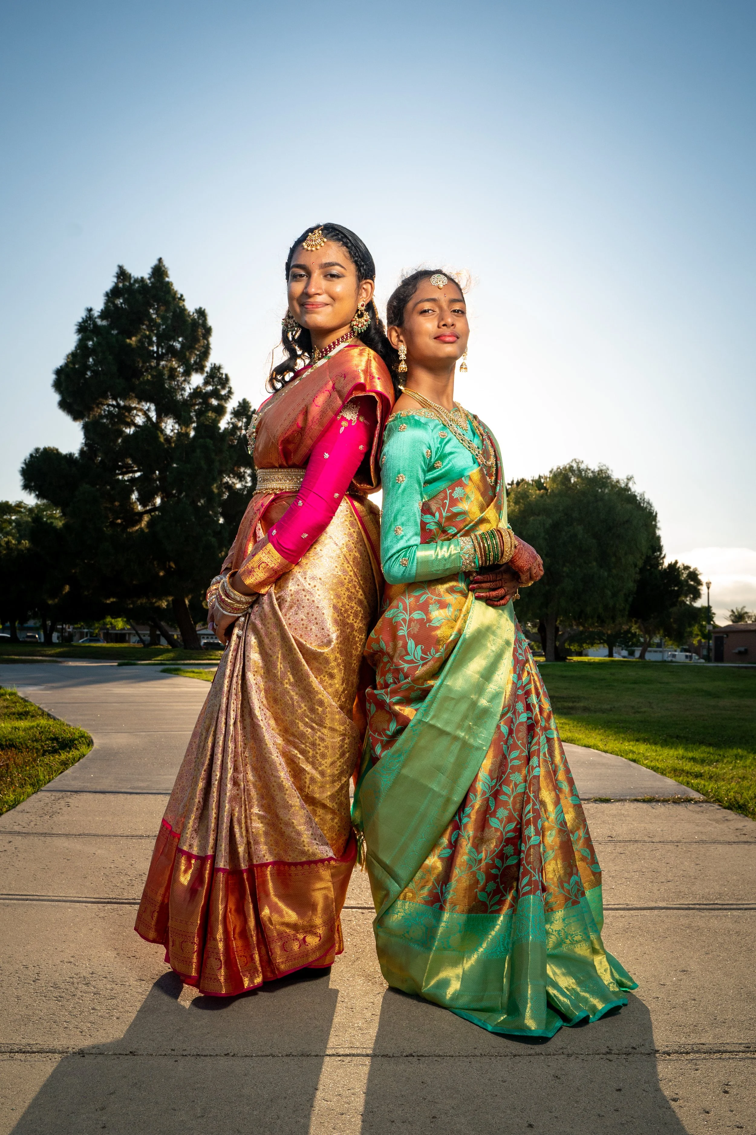 Two women dressed in traditional Indian sarees standing outdoors on a sidewalk during sunset, with trees and a park visible in the background.
