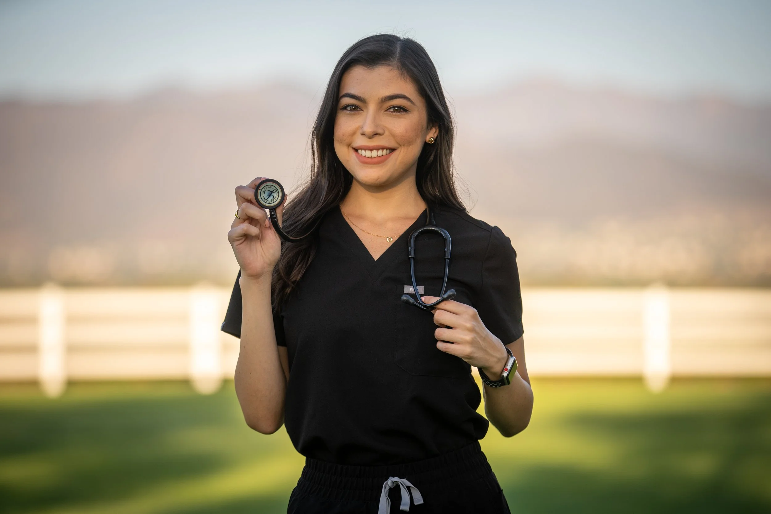A young woman in black workout attire holding a stethoscope outside during sunset, smiling at the camera.