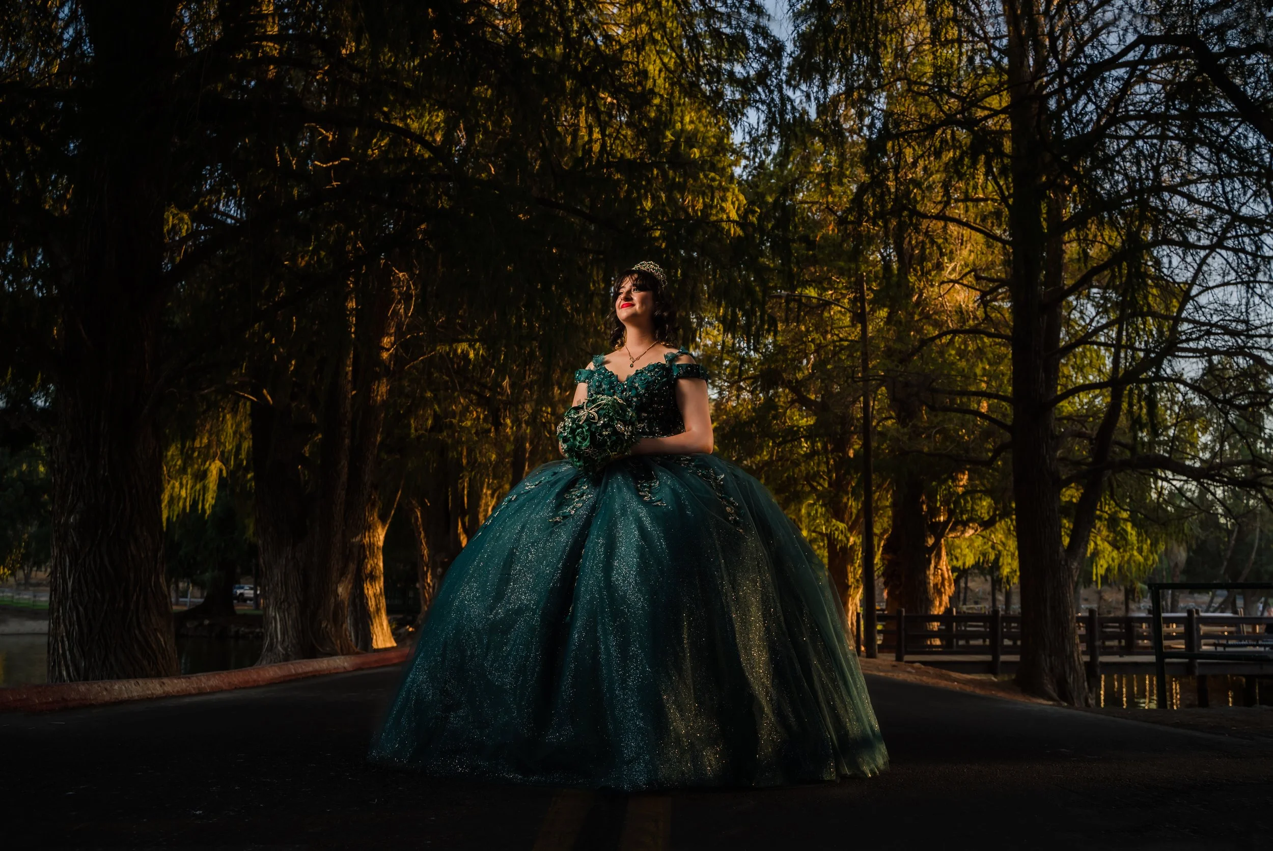 A woman in an elaborate dark green gown with floral embellishments and off-the-shoulder sleeves stands outdoors among tall trees at sunset, smiling with red lipstick and dark hair, holding a bouquet.
