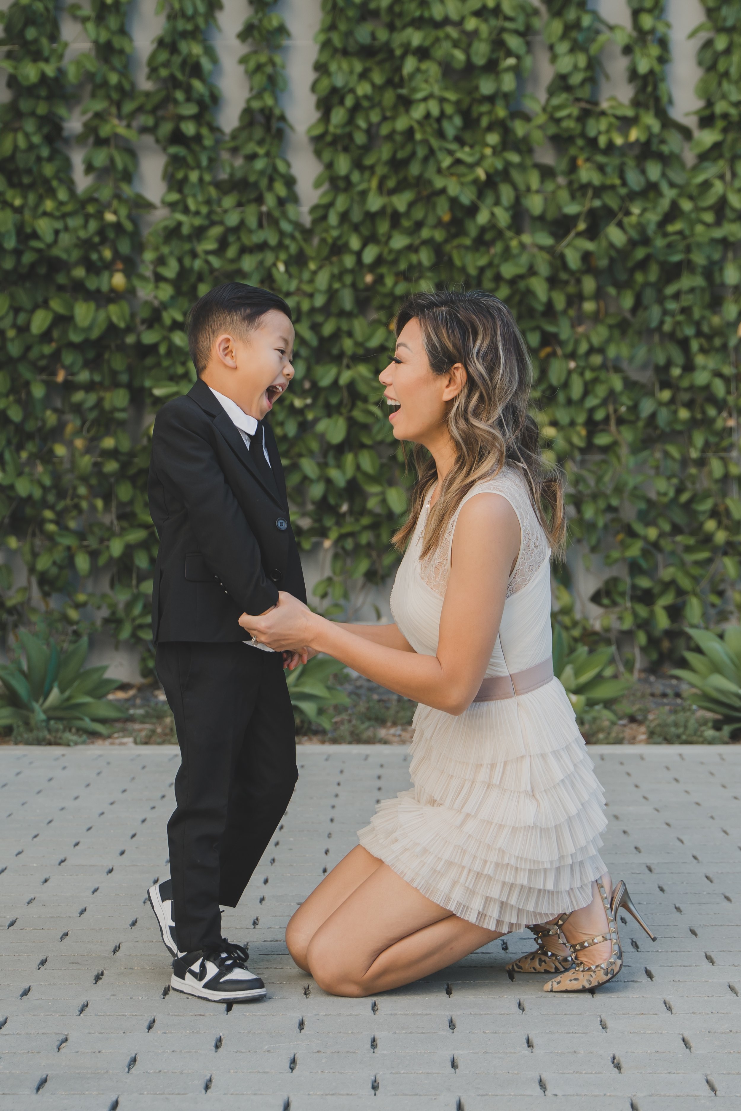 A woman kneeling on the ground, smiling, holding hands with a young boy in a suit, also smiling, in front of a green leafy background.