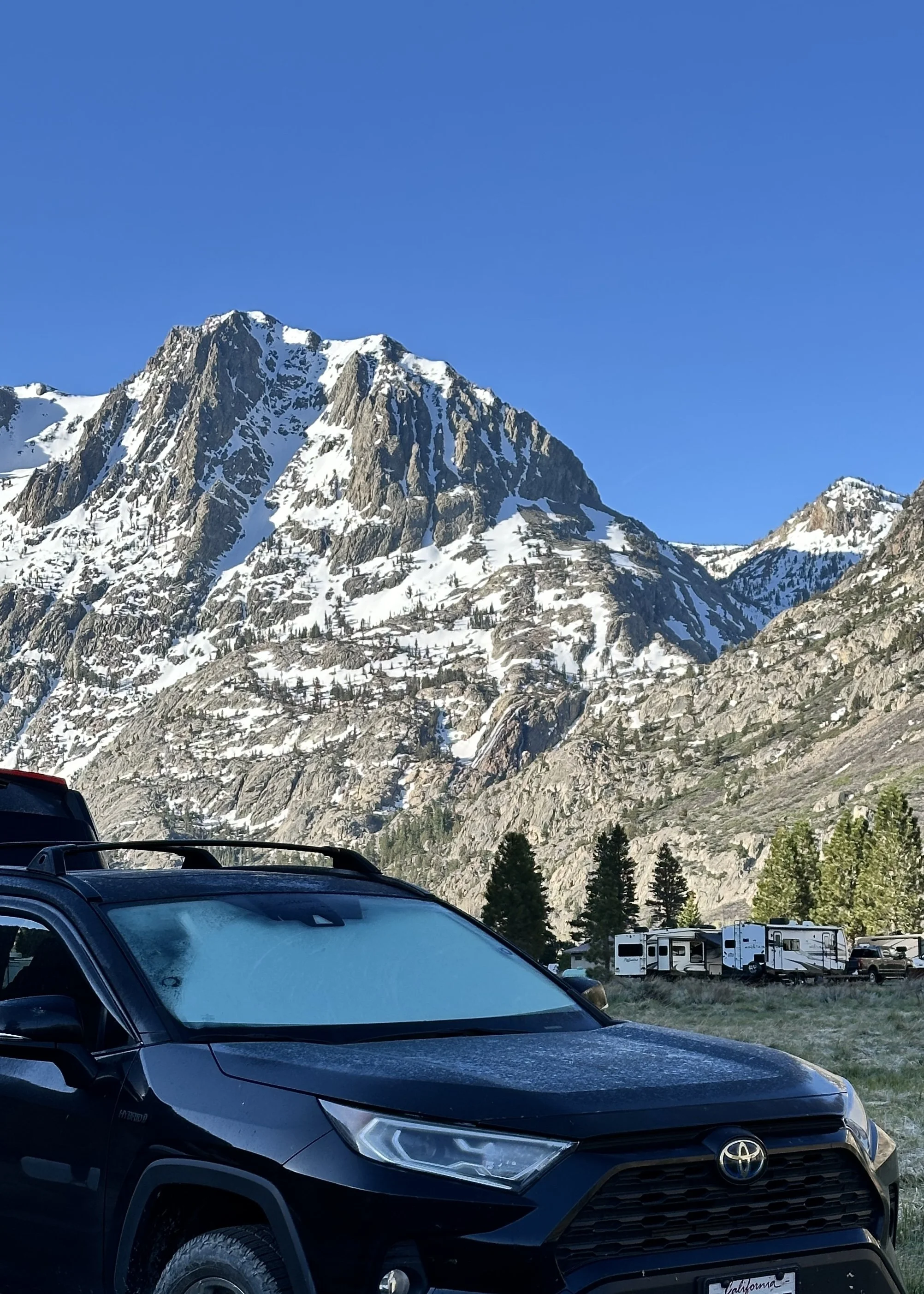 Mountain landscape with snow-covered peaks, green trees, and vehicles parked in the foreground, including a black Toyota SUV.