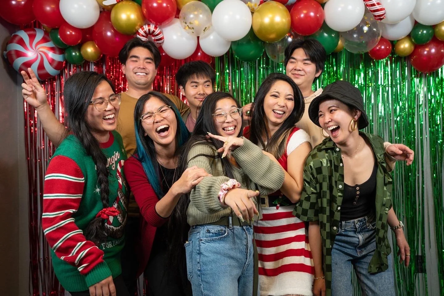 Group of friends celebrating Christmas with festive sweaters, holiday decorations, and balloons in red, green, white, gold, and clear with gold confetti, smiling and posing in front of a shiny green and red backdrop.