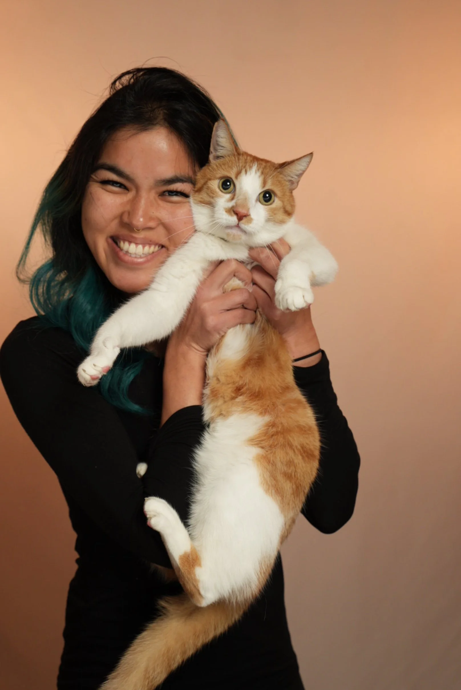 A smiling woman with black hair and teal tips is holding an orange and white cat with large green eyes, against a peach-colored background.