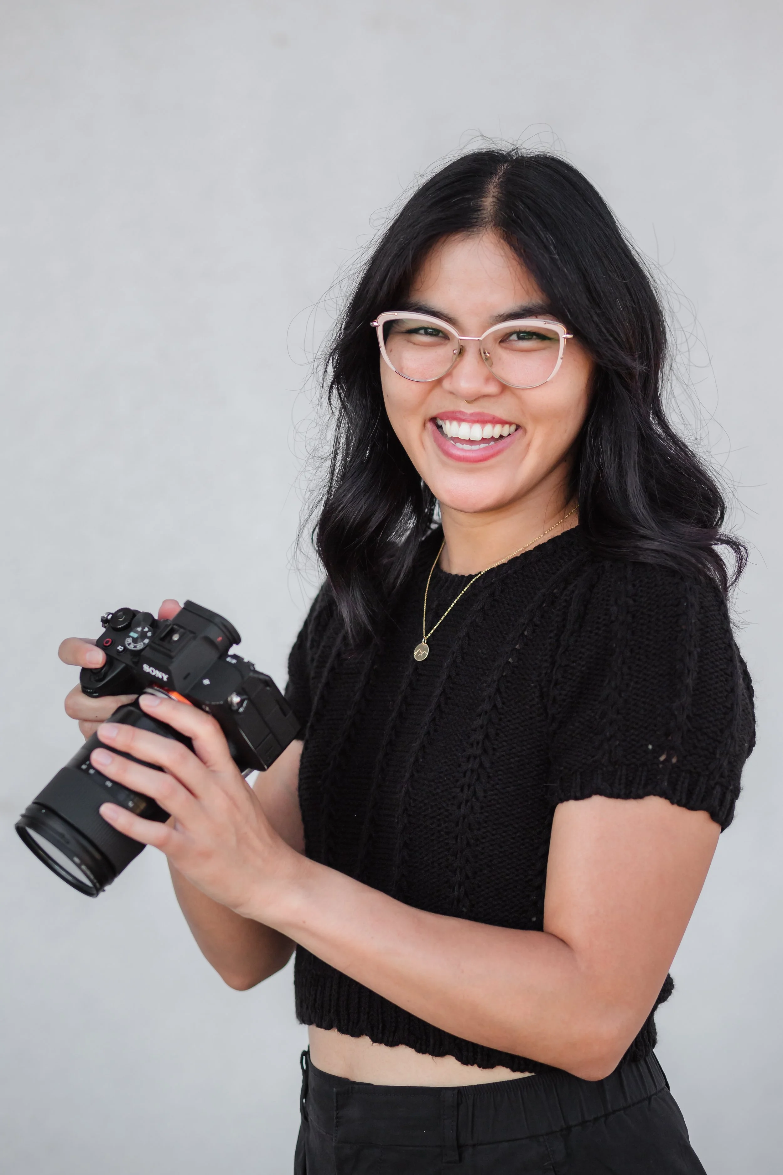 A young woman with black hair, glasses, and a bright smile, looking at the camera while holding a professional camera, wearing a black knitted top and black pants against a plain light gray background.