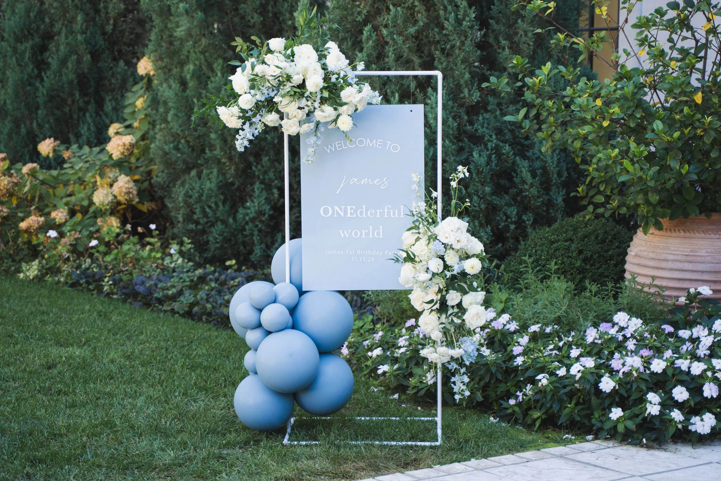 Decorative outdoor scene with a welcoming sign for a birthday party, adorned with white flowers and blue balloons, surrounded by greenery and flowering plants.
