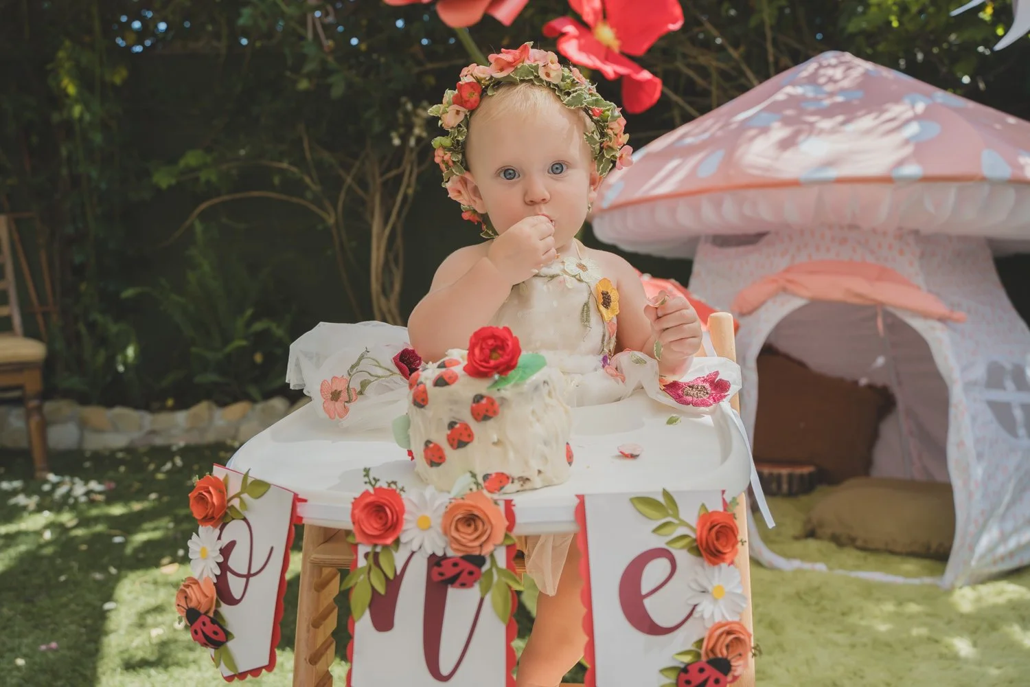 A young girl with a floral crown sitting at a decorated table with a ladybug-themed cake, celebrating her birthday outdoors with a pink and white play tent in the background.