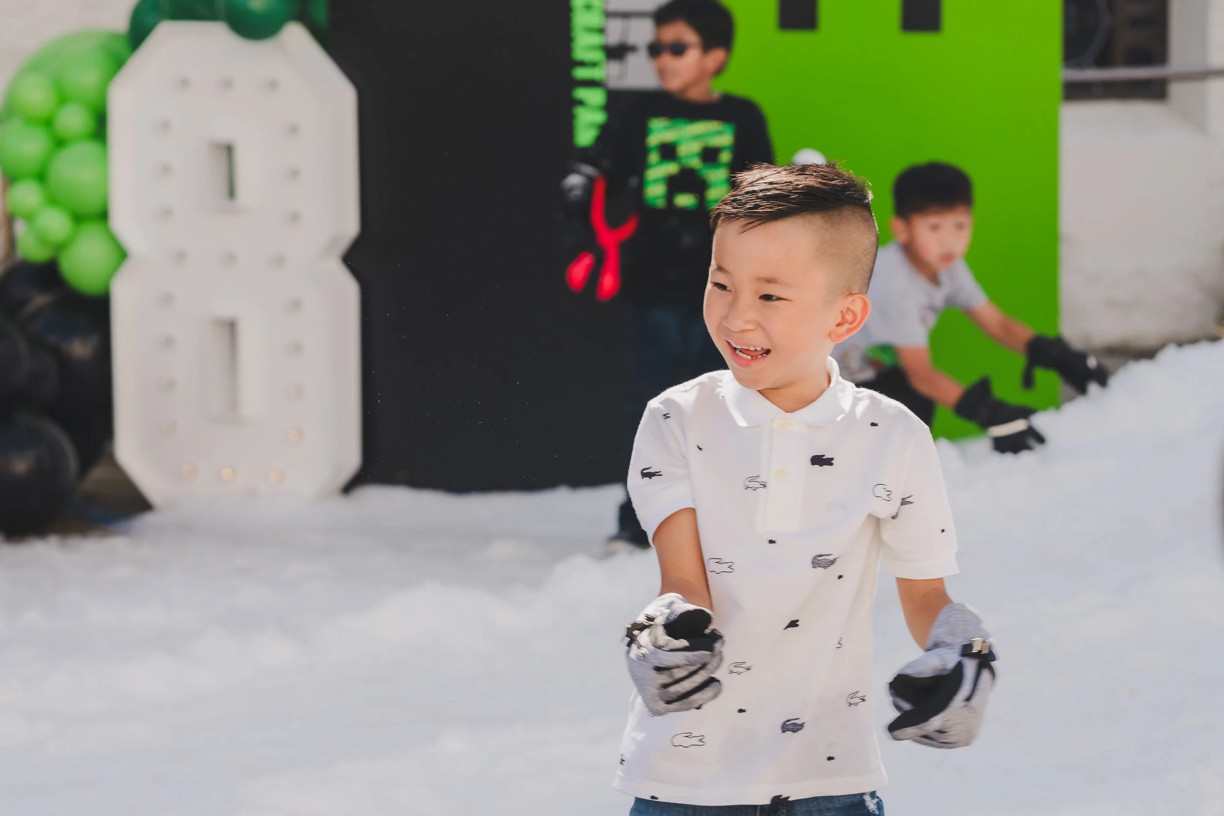 A young boy smiling and wearing a white shirt with black animal prints, standing on snow with other children and adults in the background at a winter event with green and black decorations.
