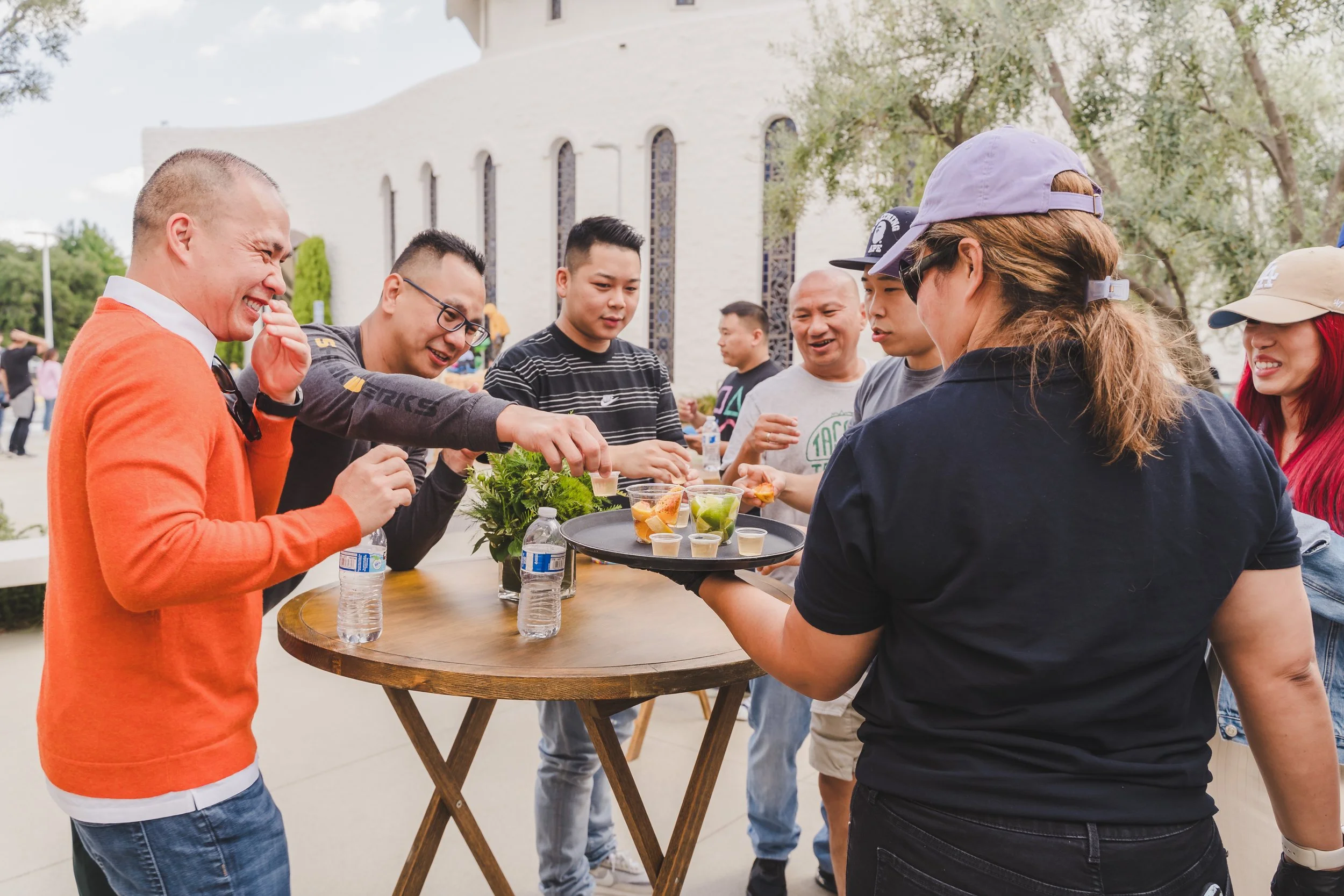 Group of people gathered around a table outdoors, enjoying drinks and conversation, with a white building and trees in the background.