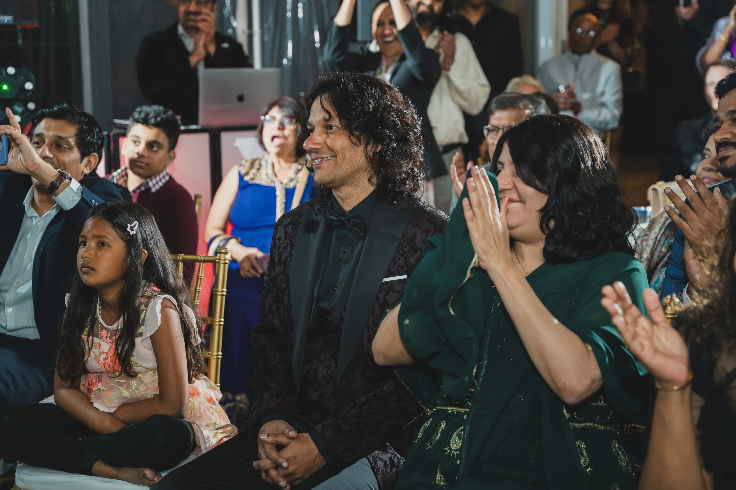 A diverse group of people attending an event, some seated and some standing, with expressions of happiness and excitement, including a smiling man in the foreground, a young girl sitting with her legs crossed, and various others clapping, taking phot