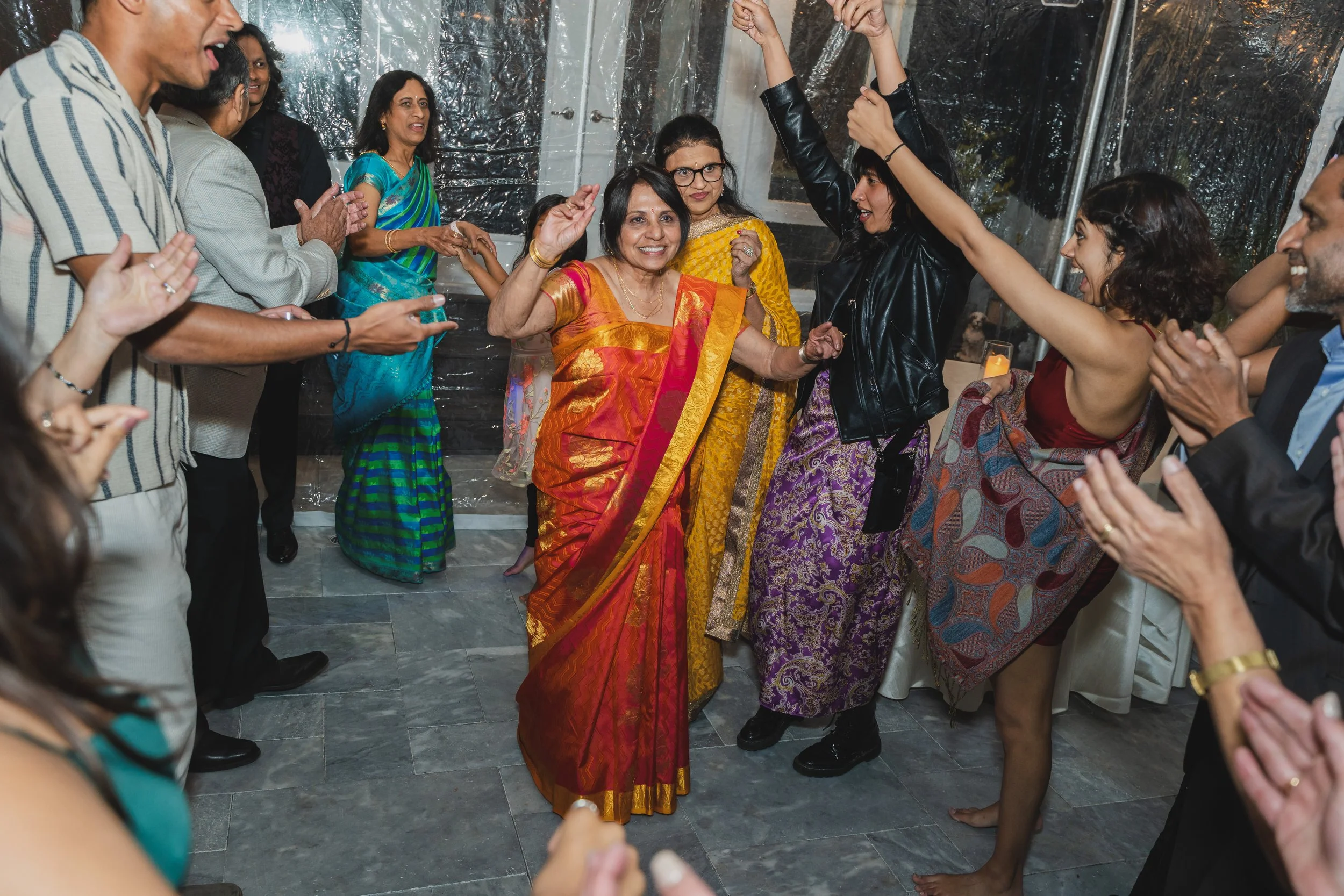 A group of people dancing and celebrating at a party, with women in colorful traditional dresses and men in casual clothes, inside a decorated venue.