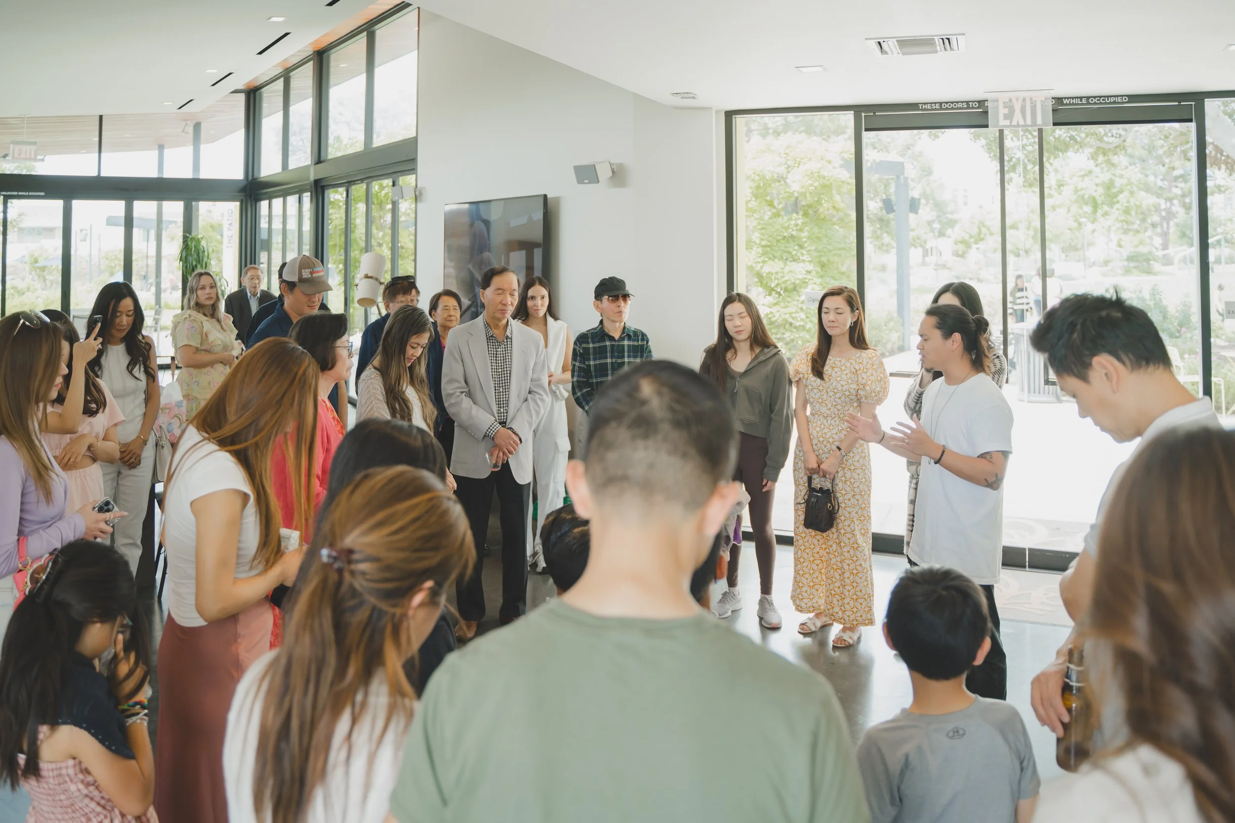 A diverse group of people gathered indoors in front of a large window, listening to a woman with tattoos speaking; the setting appears to be a modern building with glass doors and green outdoor trees.