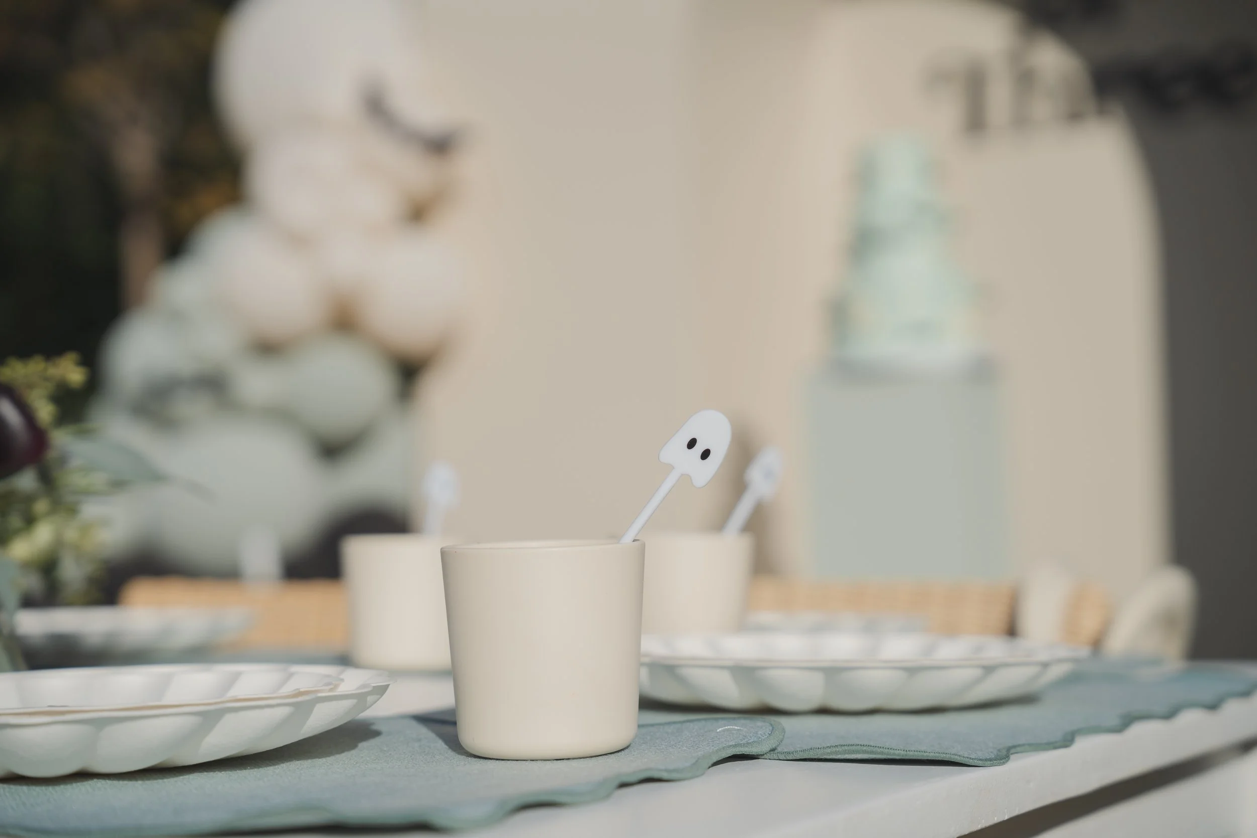 Close-up of white cups with white ghost-shaped stir sticks on a table set for Halloween or a spooky-themed event.