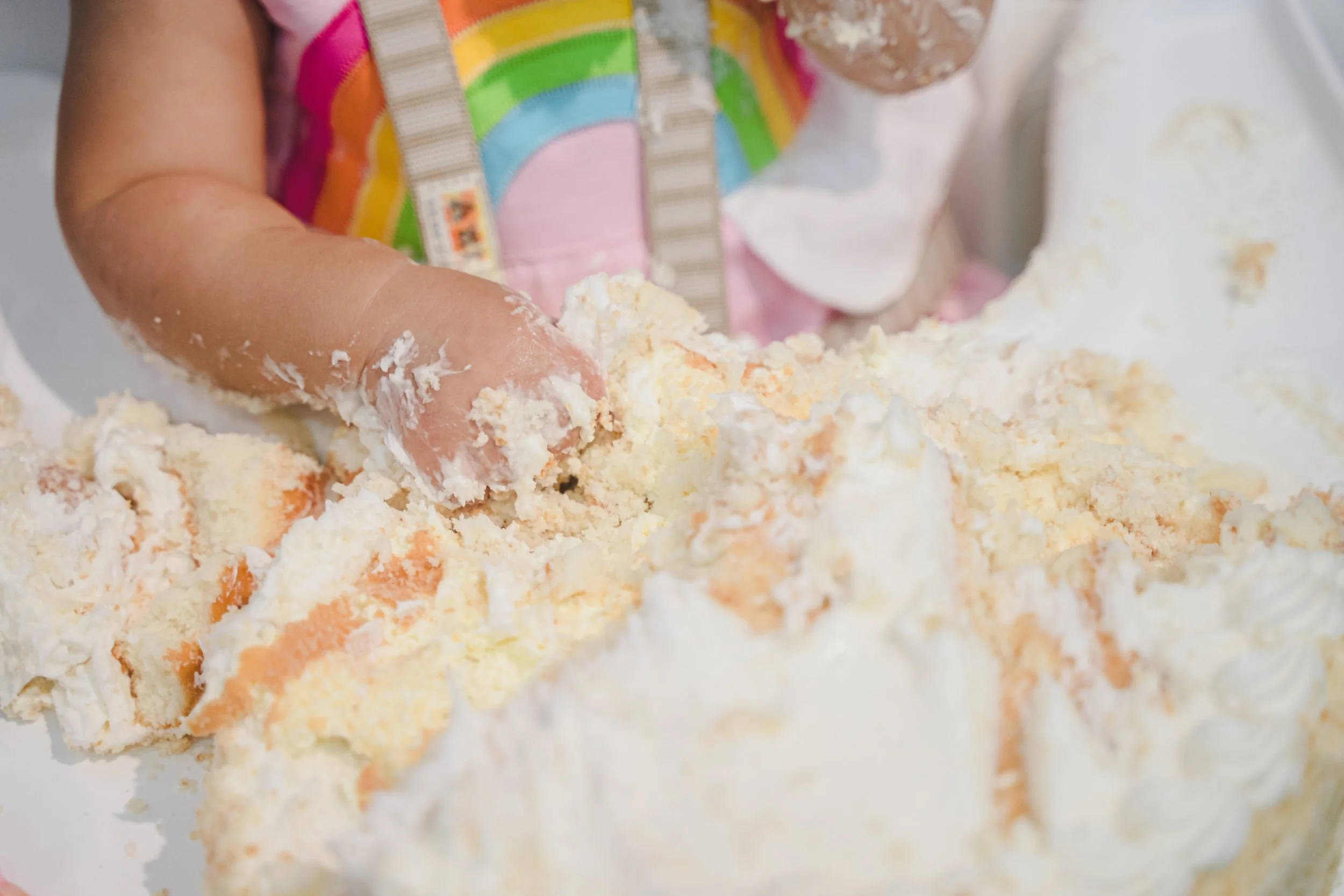 Close-up of a child's hand mixing a colorful cake batter with orange, yellow, pink, green, blue, and ivory swirls, with part of the child's rainbow-striped shirt visible.