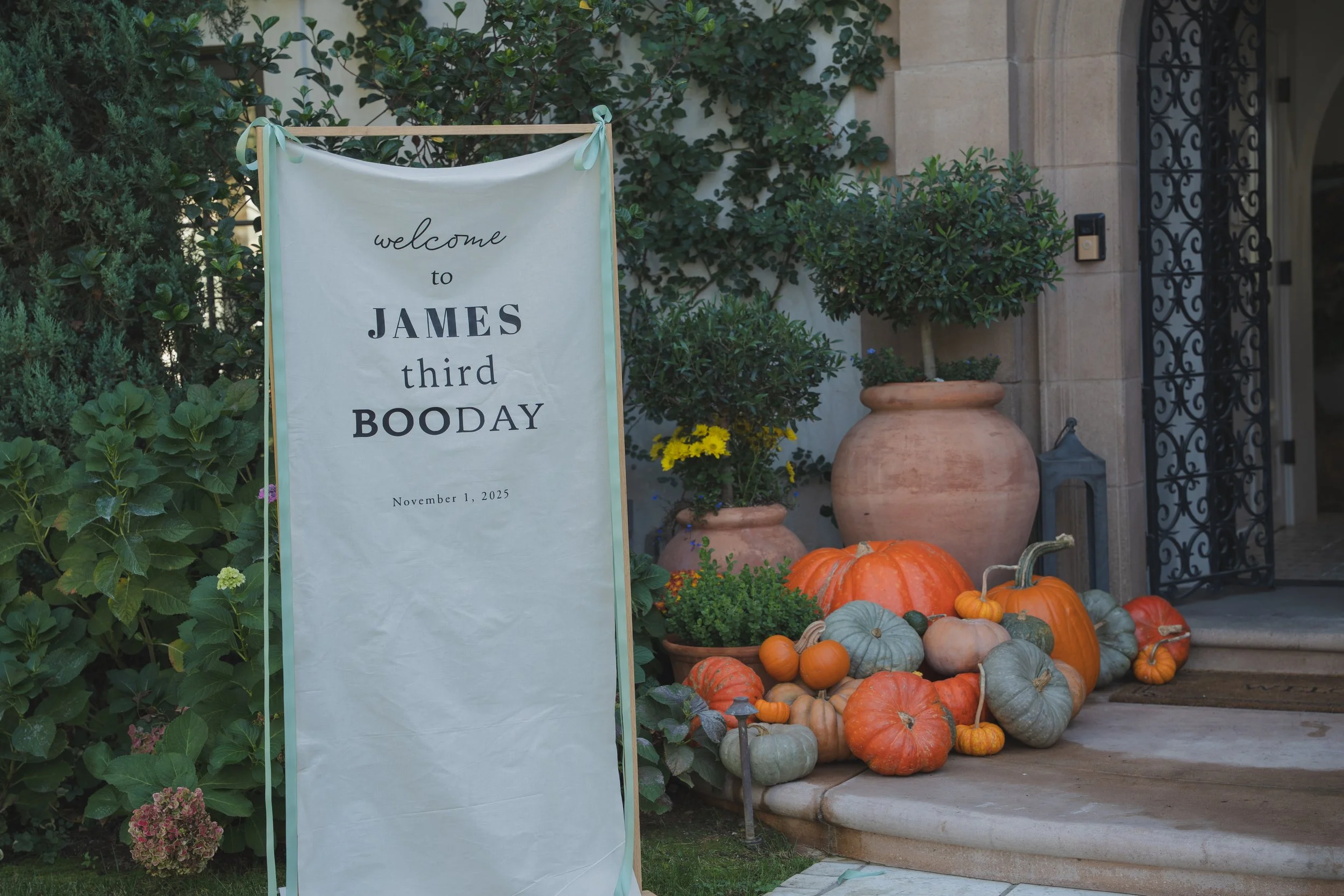 A decorative sidewalk display featuring a white fabric sign with black text that reads 'welcome to JAMES third BODAY' and the date November 1, 2025, surrounded by pumpkins and potted plants outside a building with greenery and a wrought iron gate.