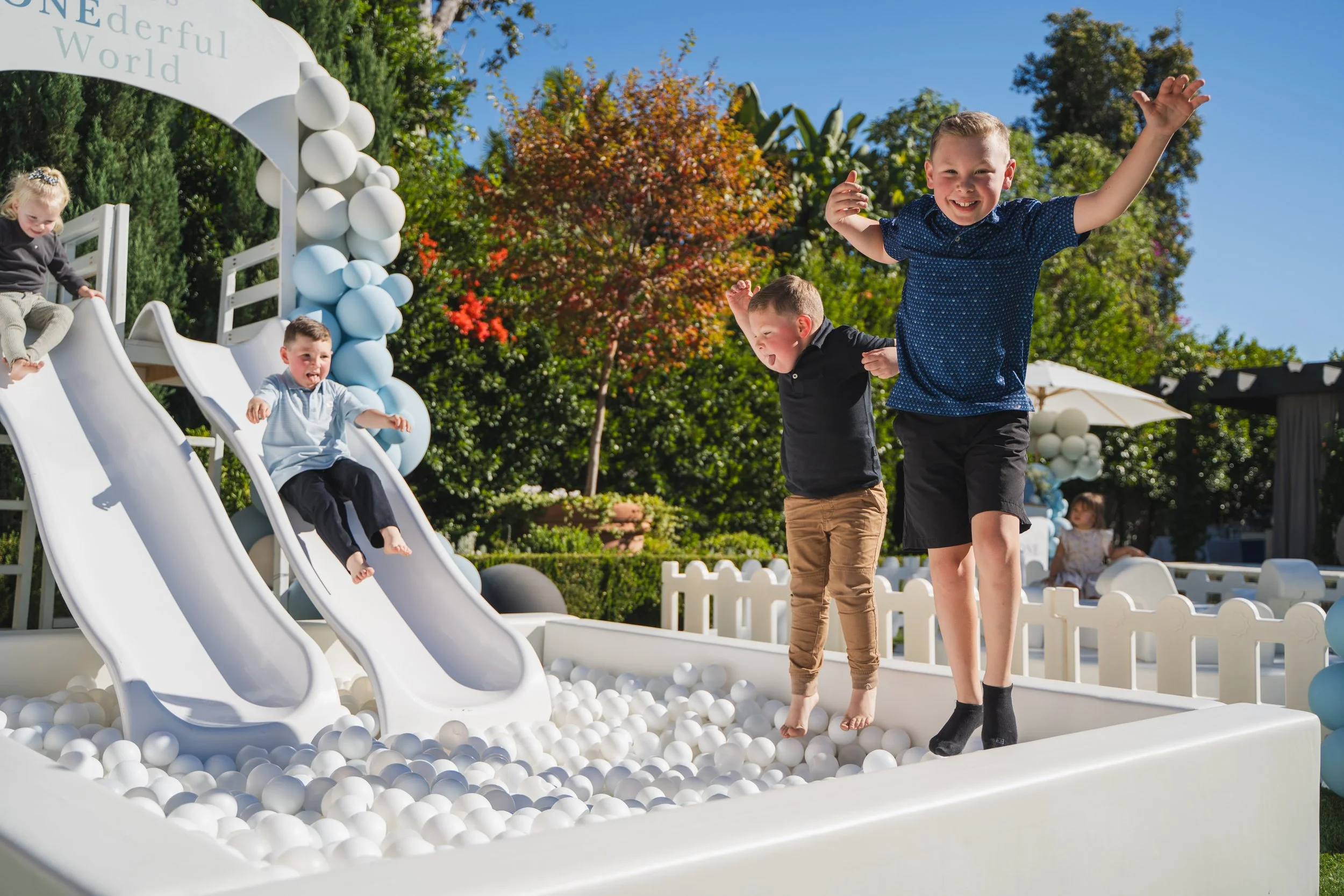 Children playing on a white ball pit with slides, surrounded by trees and greenery on a sunny day.