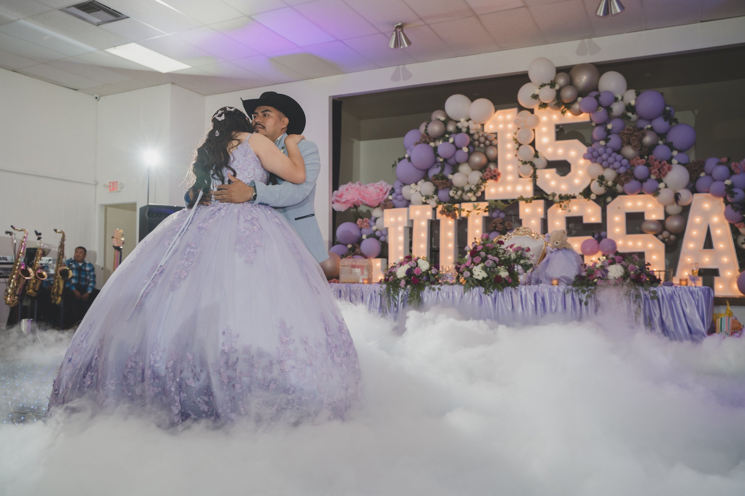Couple dancing at a wedding reception with decorations including flowers, balloons, and a large illuminated "LOVE" sign in the background.