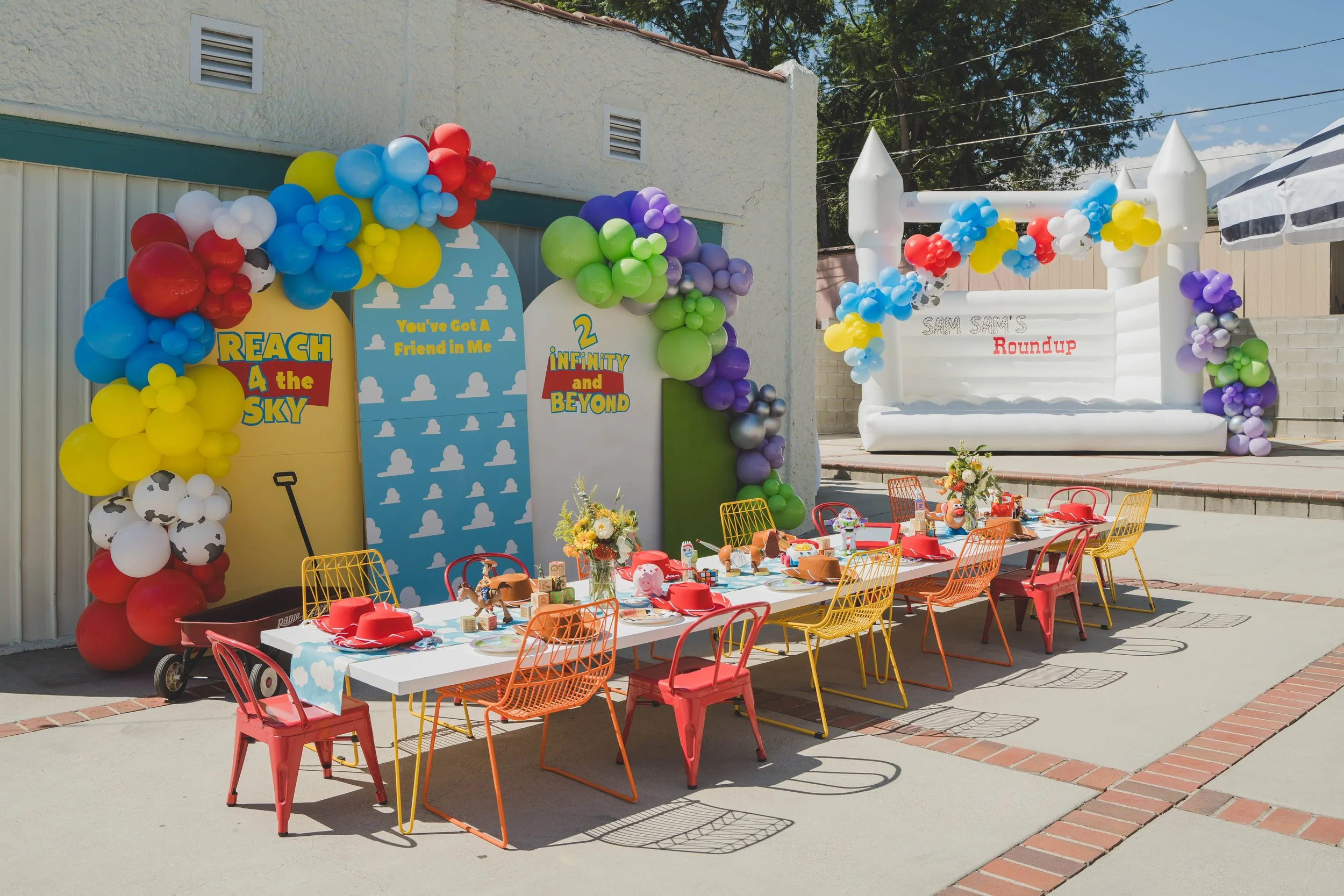Colorful outdoor party setup with tables decorated with toys, balloons, and party supplies. Balloon arches in red, yellow, blue, green, purple, and white decorate the area. A large inflatable castle with a white and black color scheme features the te