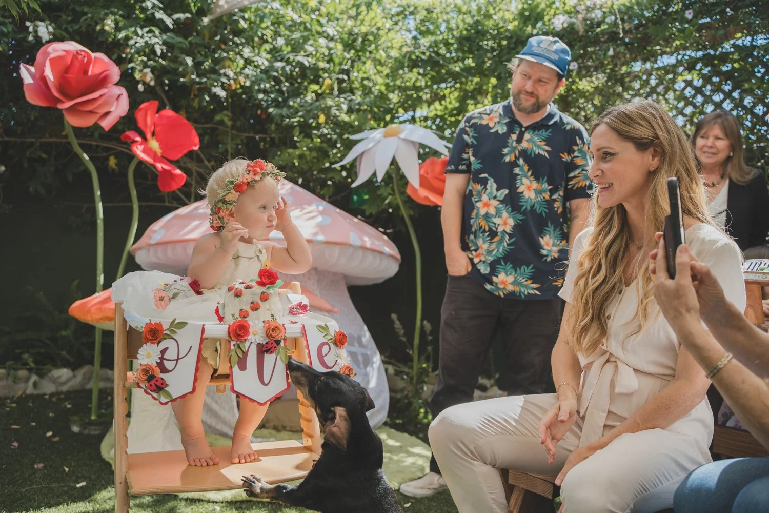 A young girl in a floral dress and crown stands on a small wooden table decorated with flowers and ladybug cutouts, touching her face during a birthday celebration outdoors. An adult woman with long blonde hair, dressed in white, sits nearby, smiling