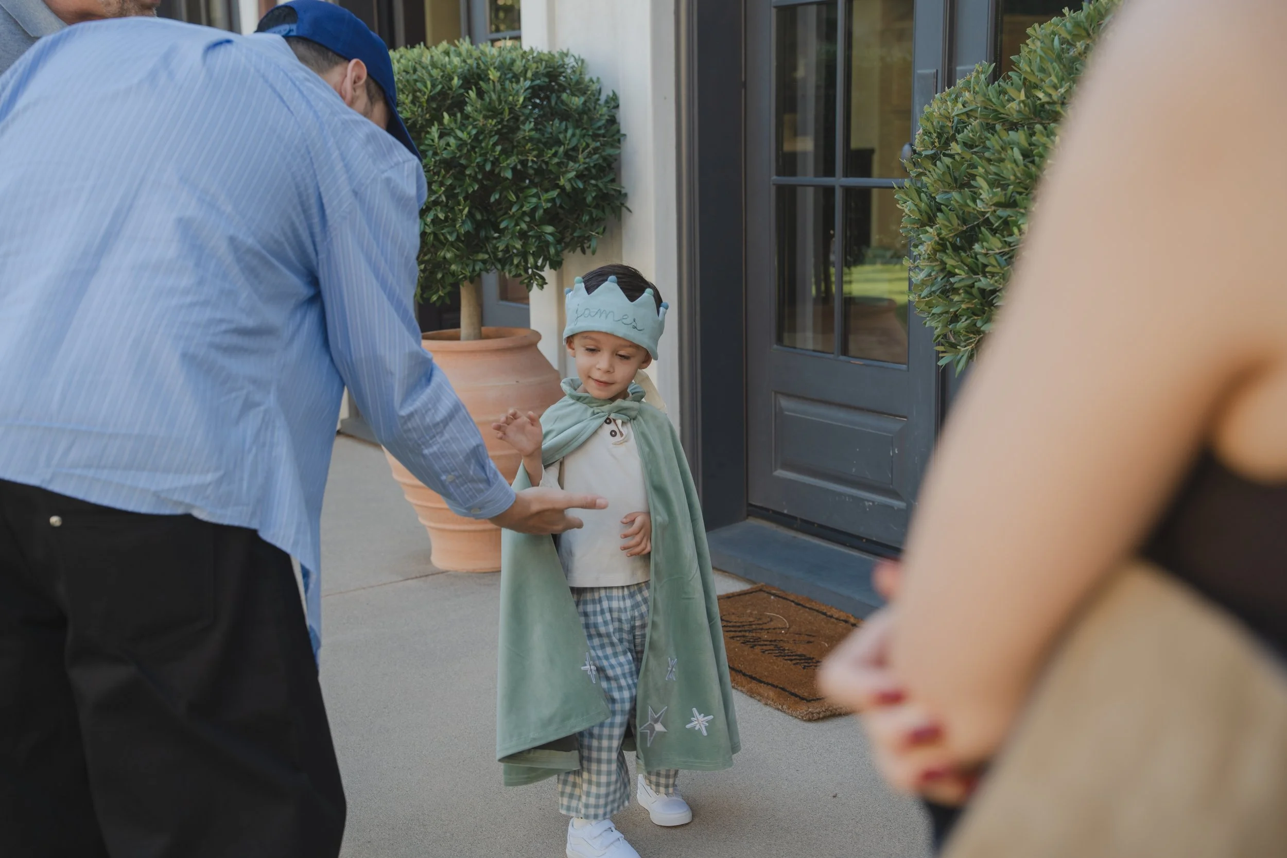 A young boy dressed as a king with a paper crown, standing outside a house, waving at someone, with adults around him.