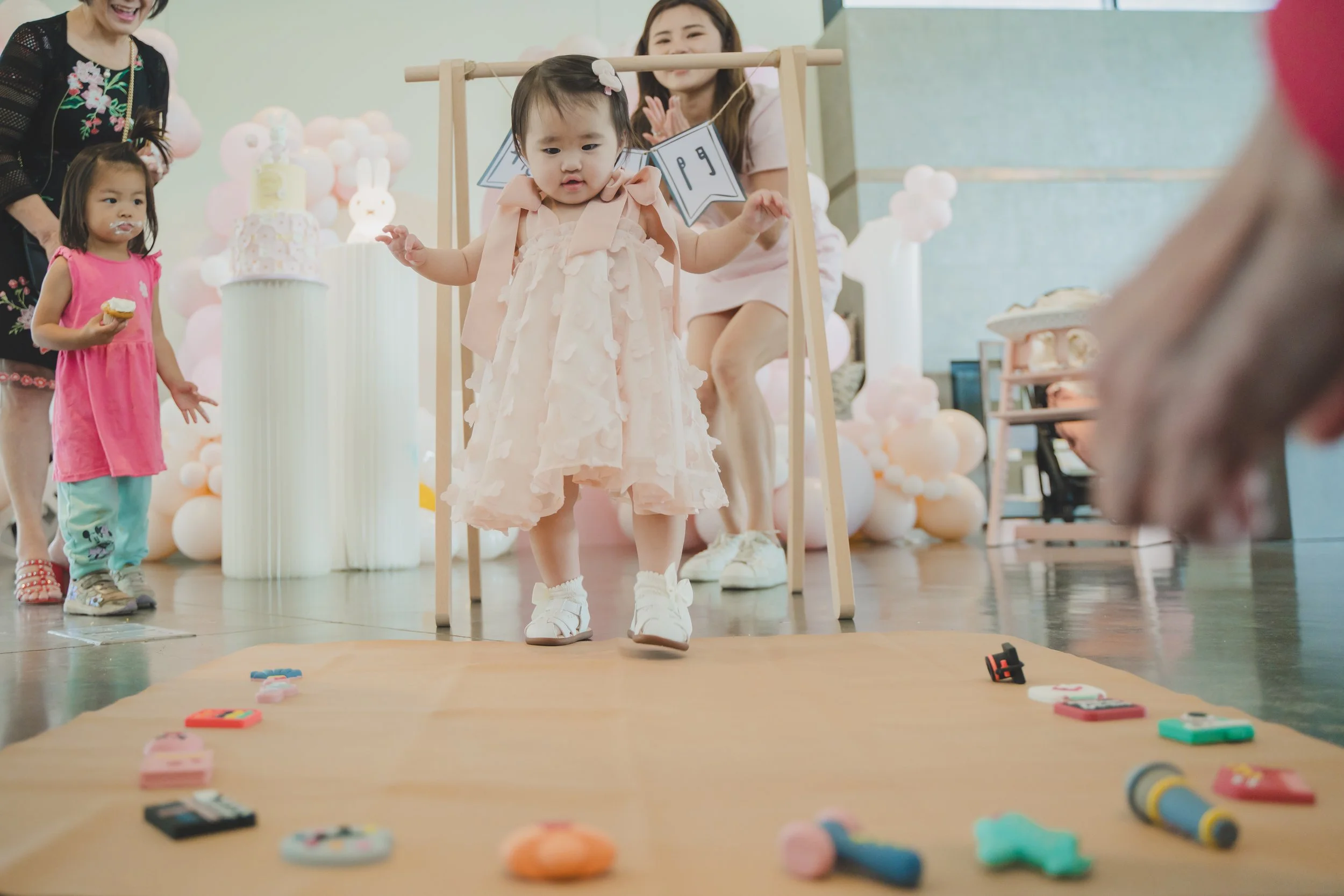 A young girl in a pink dress is standing on a wooden floor holding a small item, with a group of people and decorations behind her, including a multi-tiered cake and pink balloons, suggesting a celebration or party.
