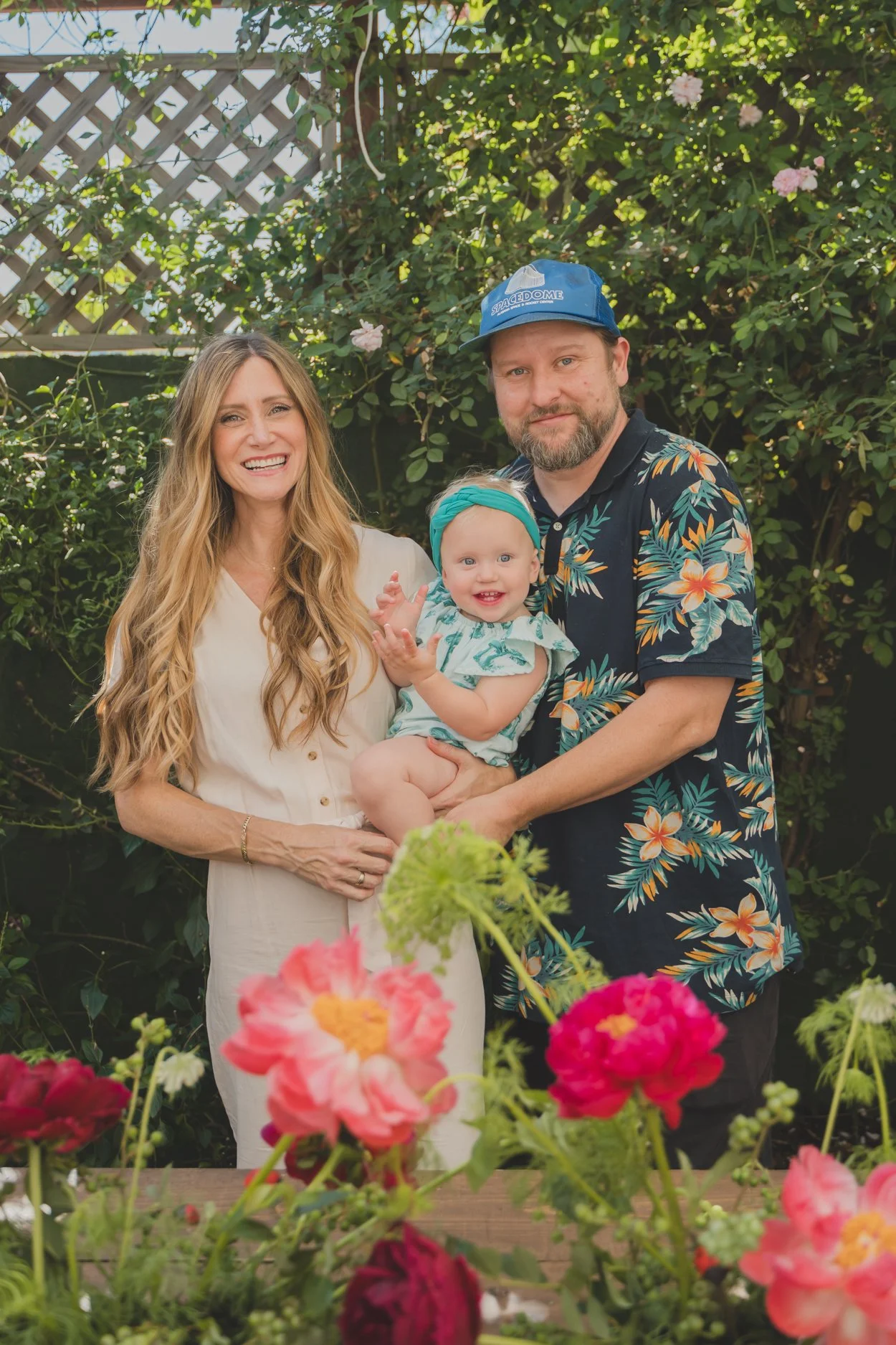 A family of three standing outdoors in a garden filled with pink, red, and green flowers and green foliage. The woman has long wavy blonde hair and is wearing a beige dress. The man has a beard, wearing a blue cap and a black Hawaiian shirt with colo