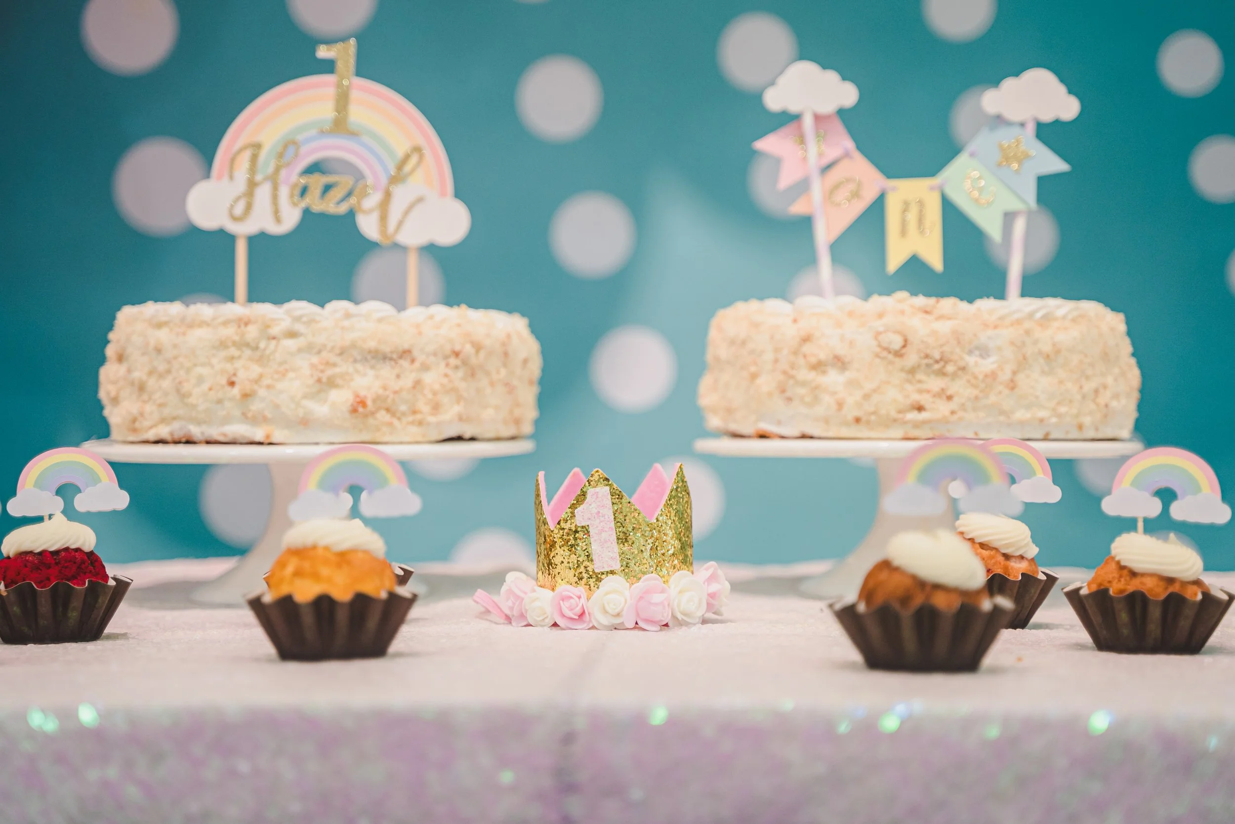 Decorative birthday cake display with cupcakes, a cake with a rainbow topper, and birthday banners on a pastel blue background with white polka dots.