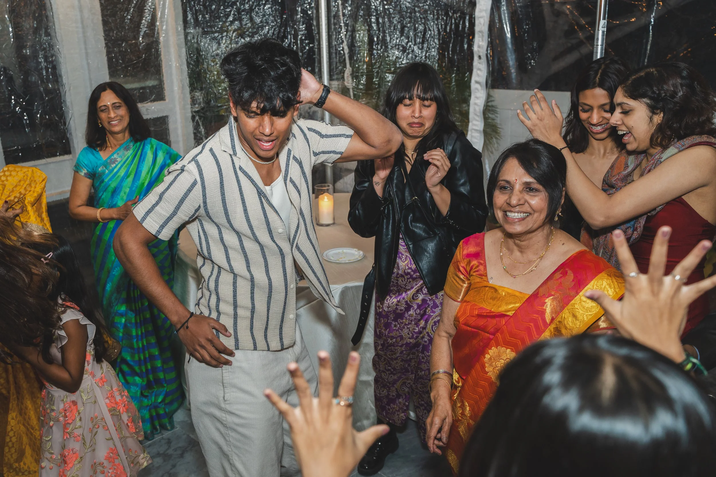 A group of people, mainly women wearing colorful traditional Indian clothing, celebrating and dancing together with happy expressions indoors at a festive gathering.