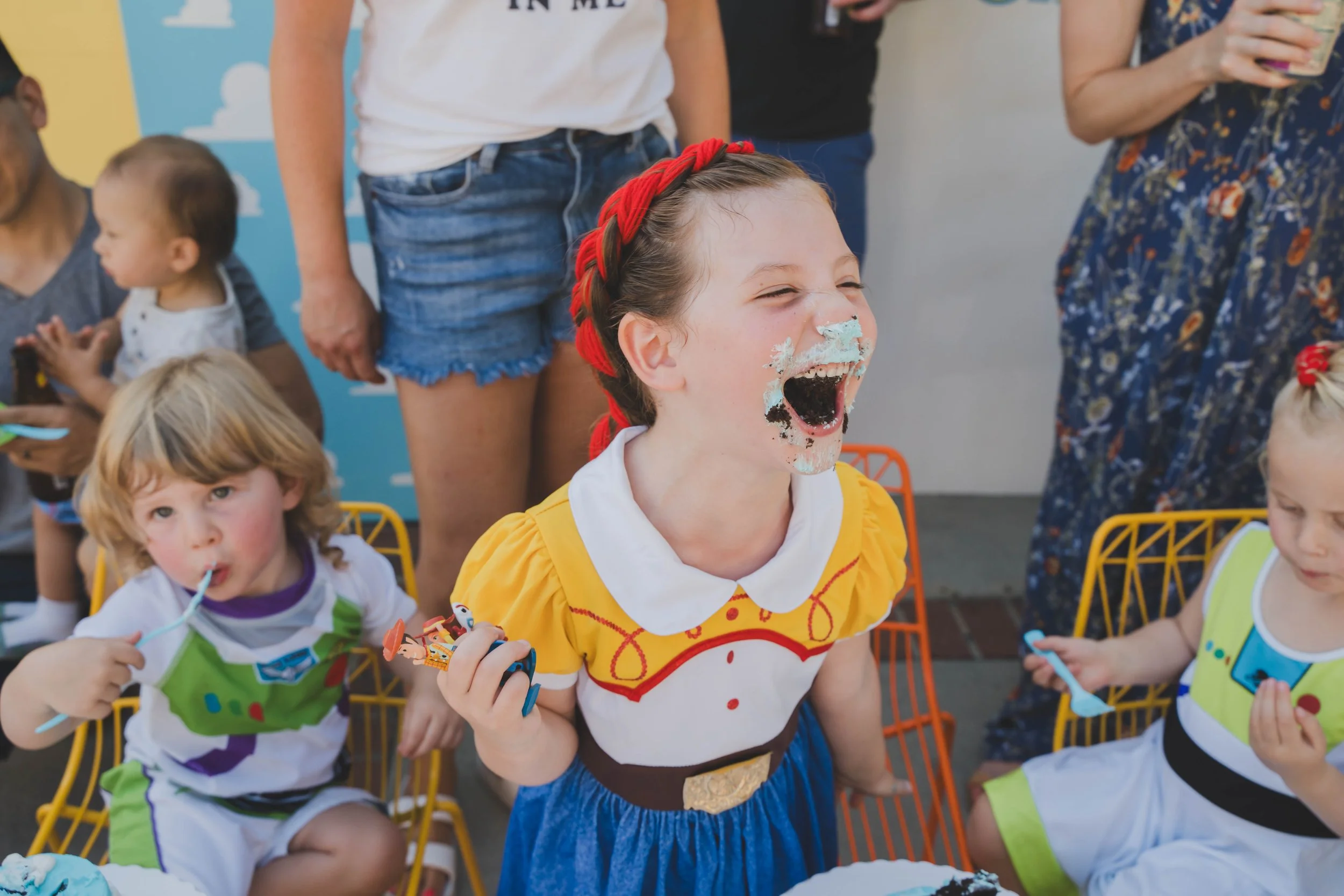 Children celebrating birthday, girl with face smeared with cake, wearing yellow dress with red headband, surrounded by others at party.