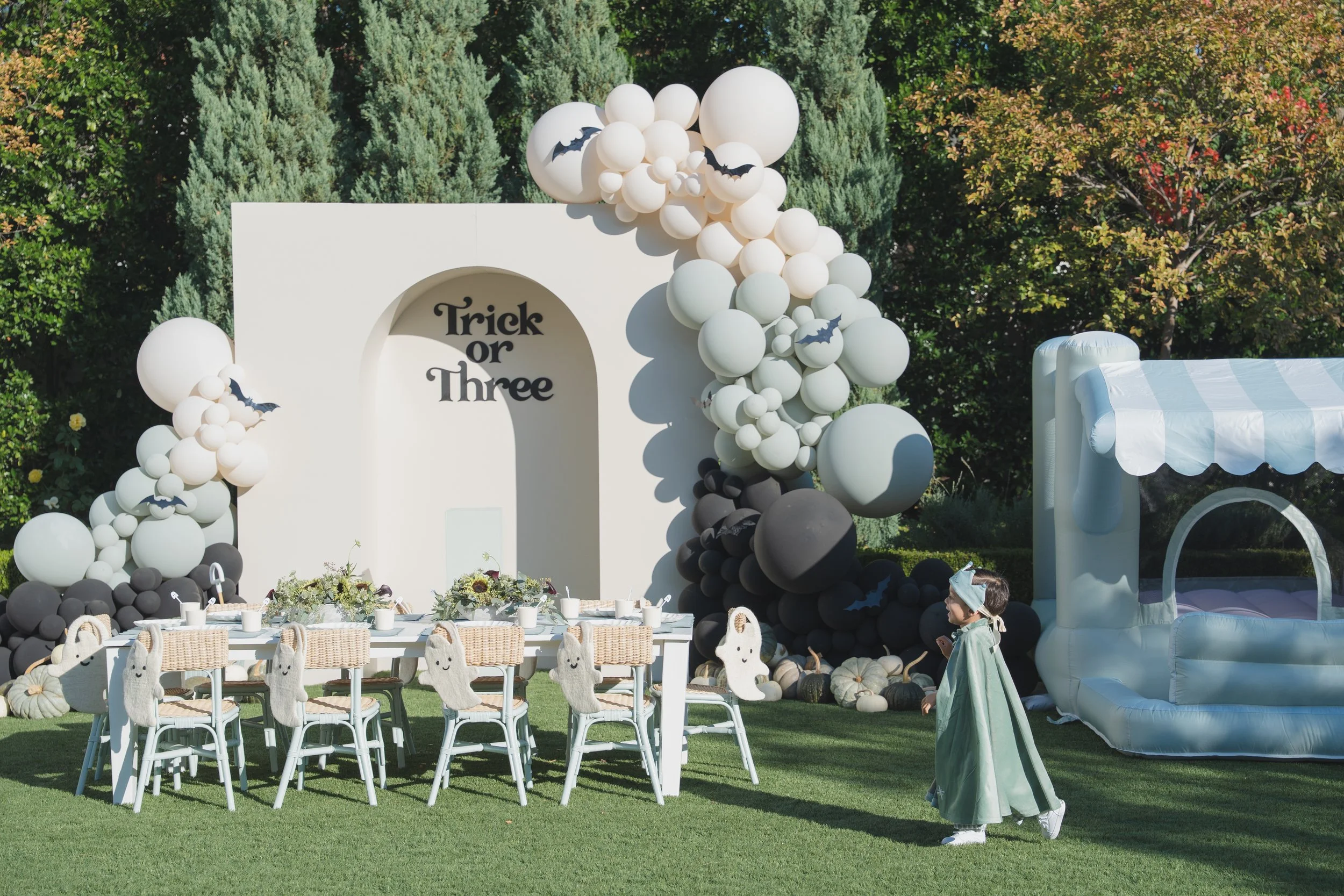 Outdoor Halloween-themed party setup with white and black balloons, some with bat shapes, a white arch with 'Trick or Three' written on it, a table with ghost decorations on the chairs, and a large inflatable structure resembling a candy or treat bag