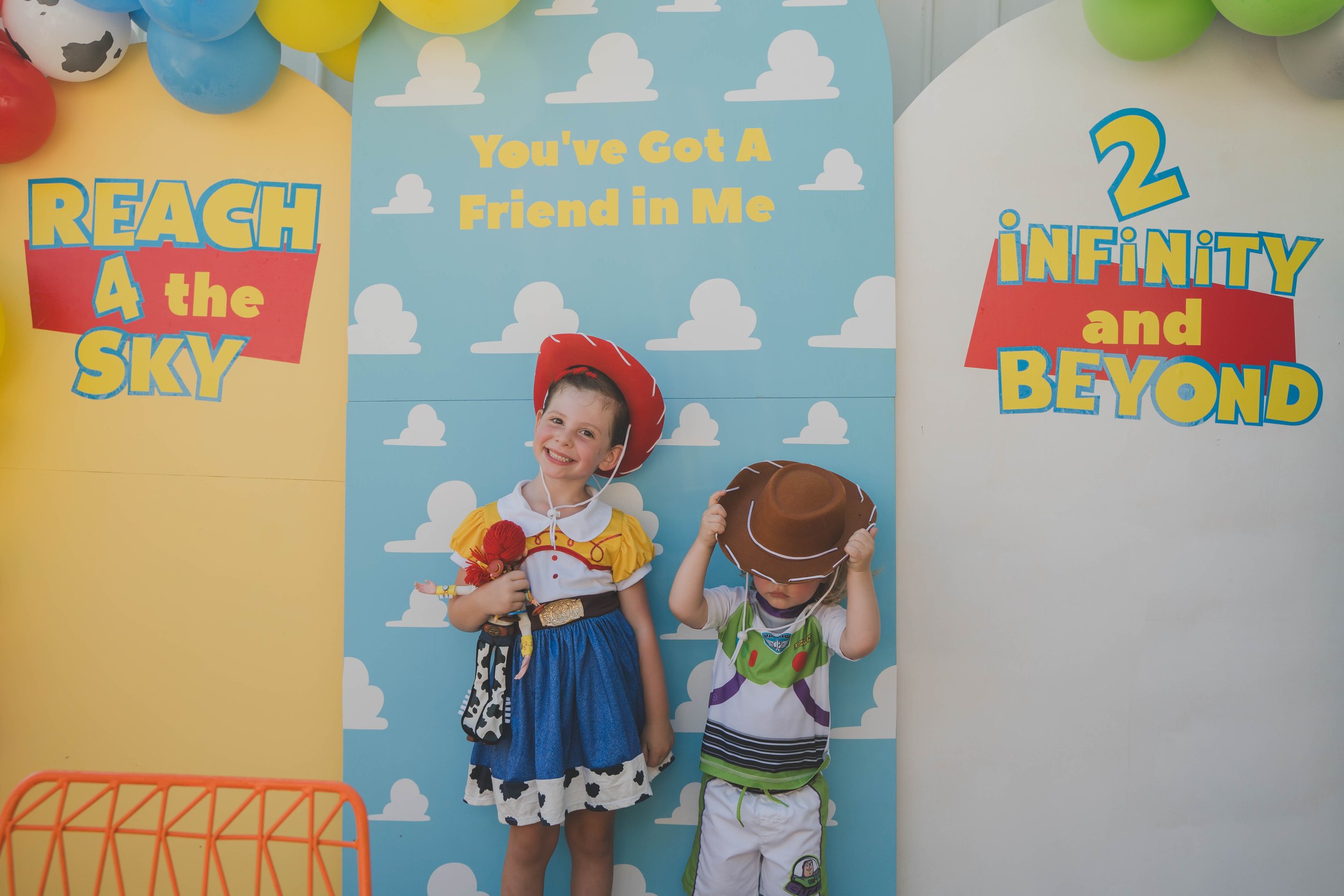 Two children dressed in costumes are standing in front of a colorful backdrop with sky and clouds. The girl is smiling and wearing a cowgirl outfit with a red cowboy hat, and the boy is wearing a costume with a hat that looks like a chocolate donut a