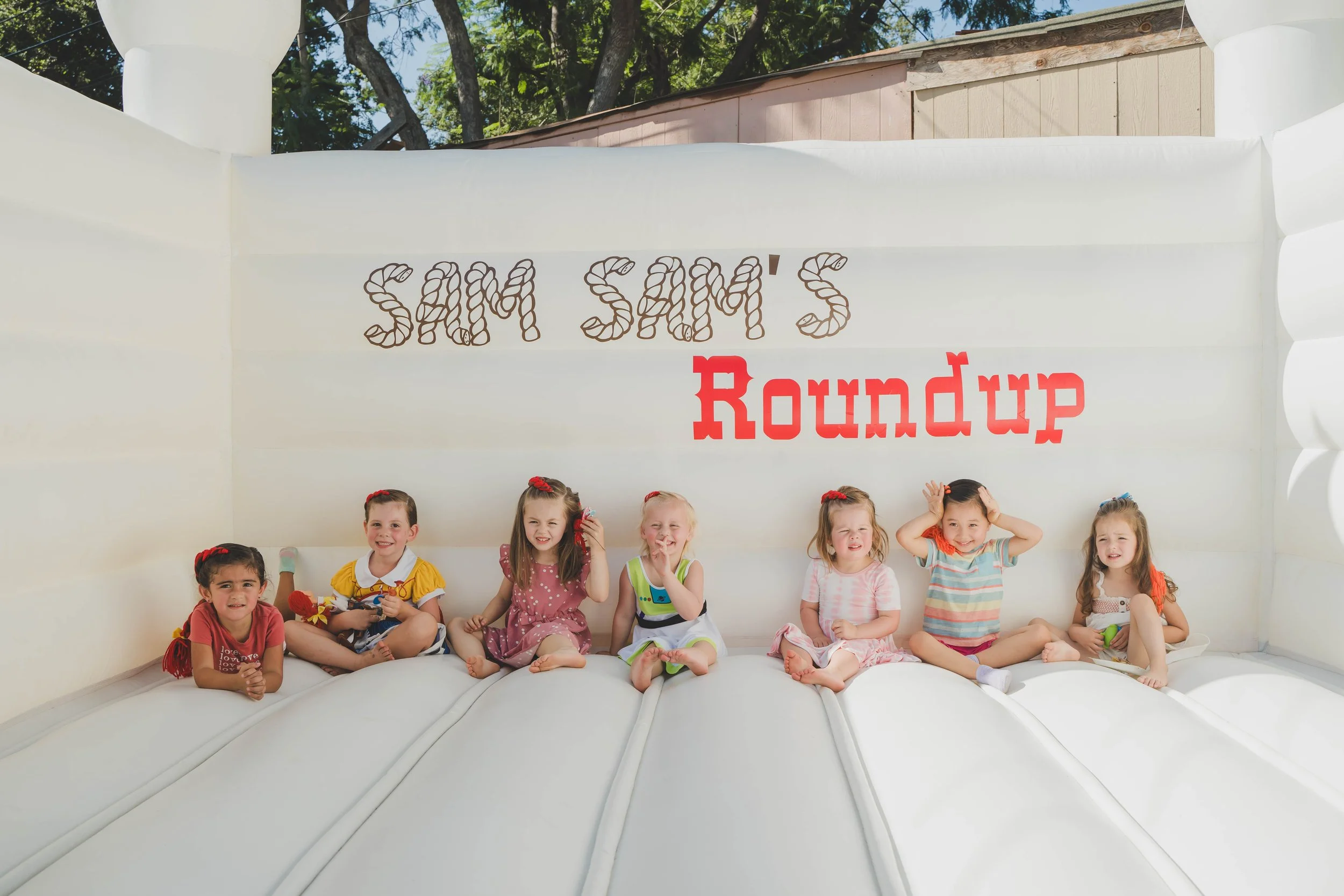 A group of young children sitting on a white inflatable bounce house with a large sign that reads 'Sam Sam's Roundup' in the background, with trees and a building visible behind the bounce house.