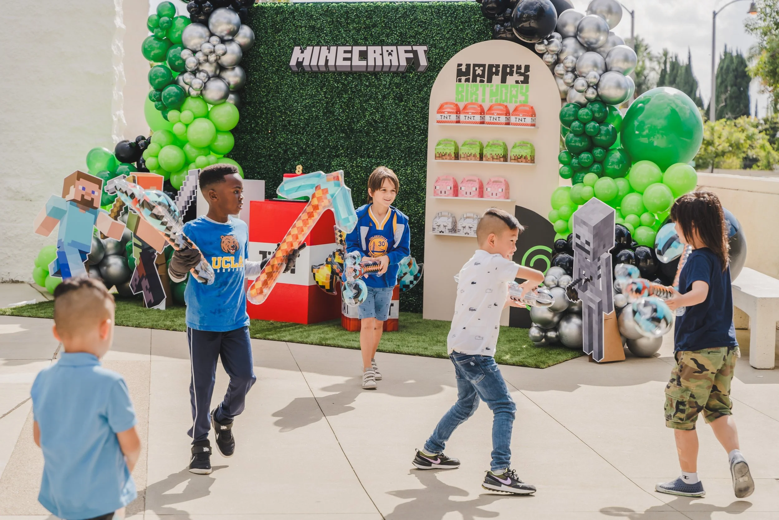 Children celebrating a Minecraft-themed birthday party with a decorated backdrop, balloons, and Minecraft cardboard cutouts.
