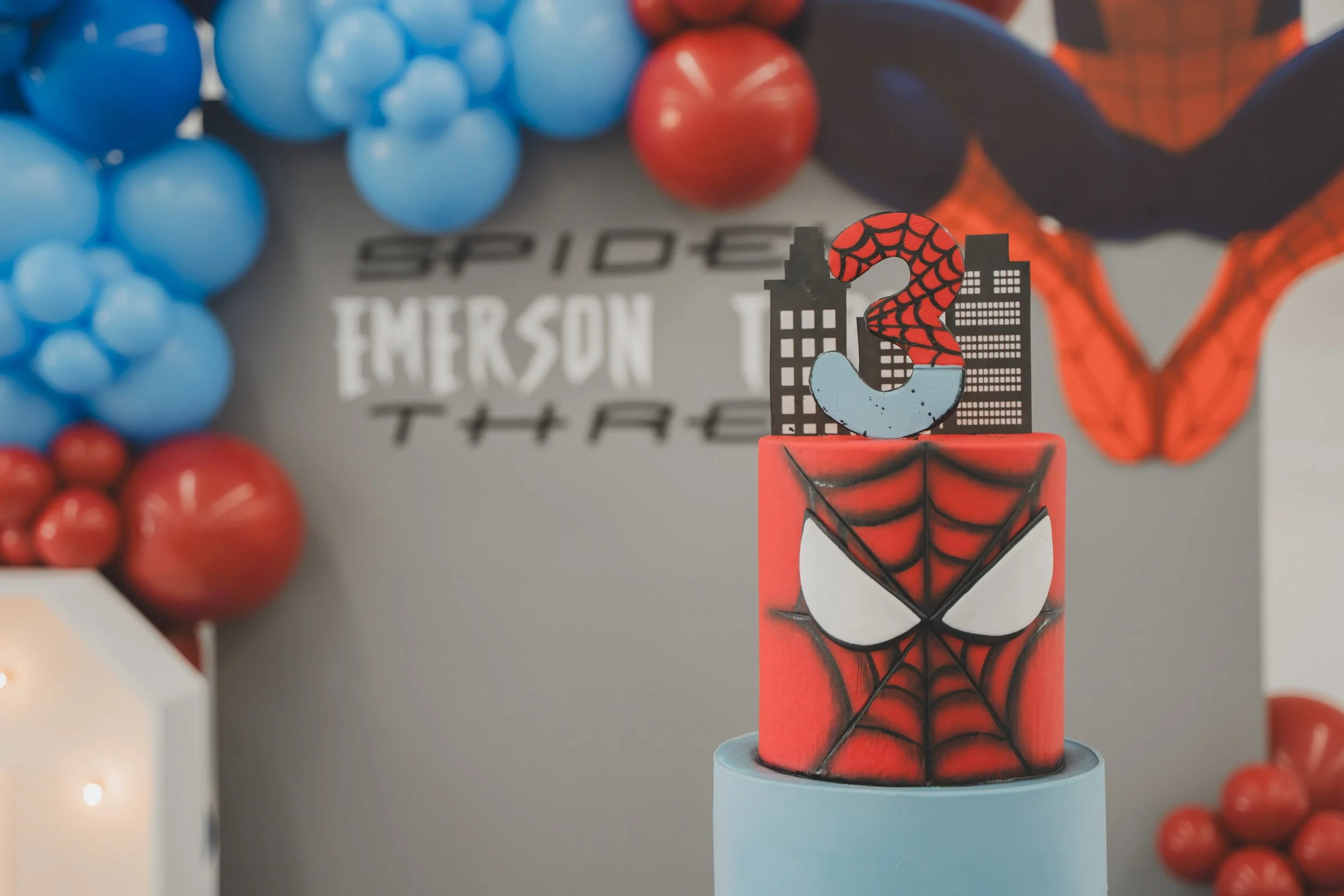 A cake decorated with a Spider-Man theme, featuring a Spider-Man mask and web design, with a number 3 topper in red and black. Blue and red balloons in the background, and a wall with Spider-Man artwork.
