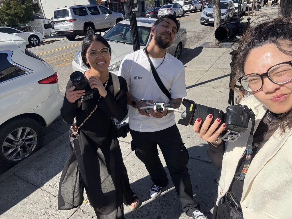 Three smiling people holding cameras on a city sidewalk, with parked cars and street in the background.