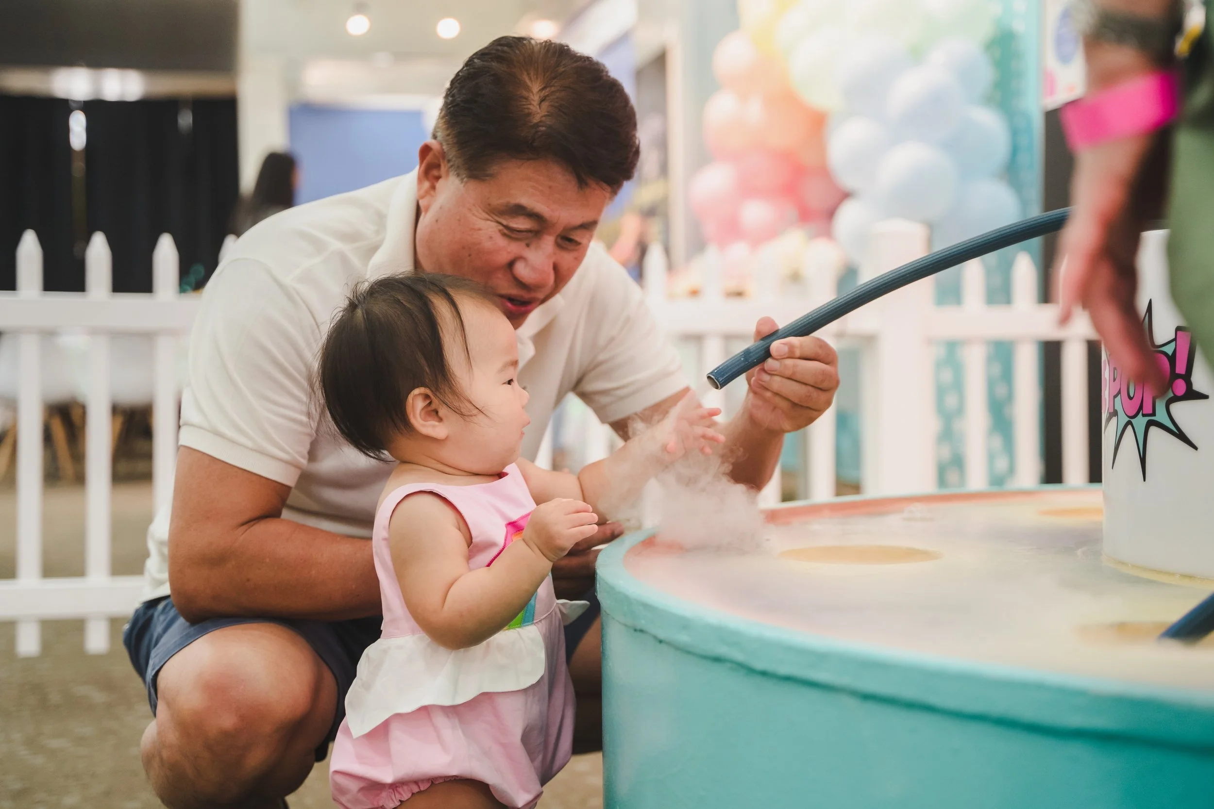 A man and a young girl are playing a carnival game with cotton candy on a colorful counter, surrounded by balloons and festive decorations.