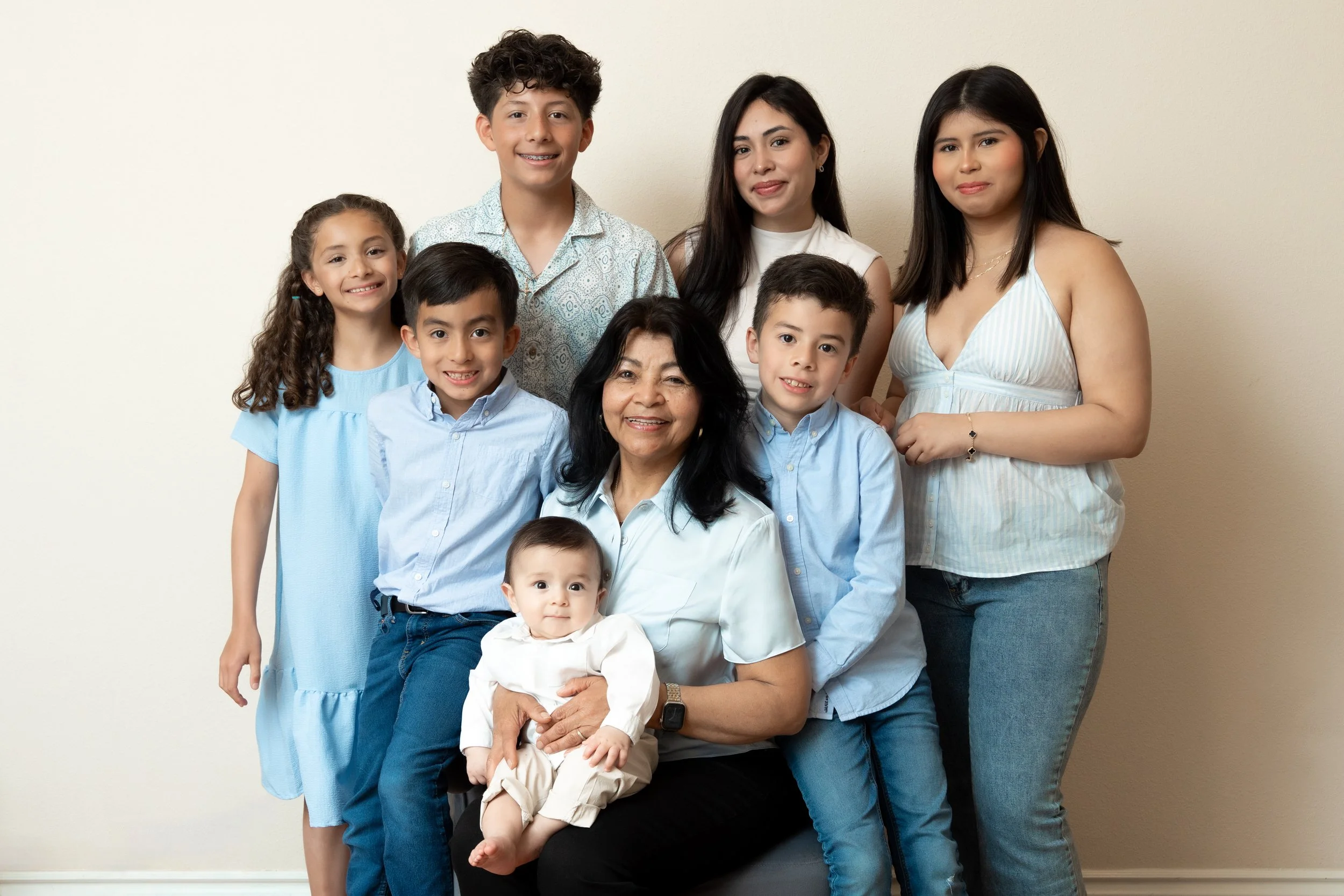 A diverse multi-generational family portrait against a plain beige wall, featuring six children, two women, and one elderly woman, all smiling and dressed in casual, colorful clothing.