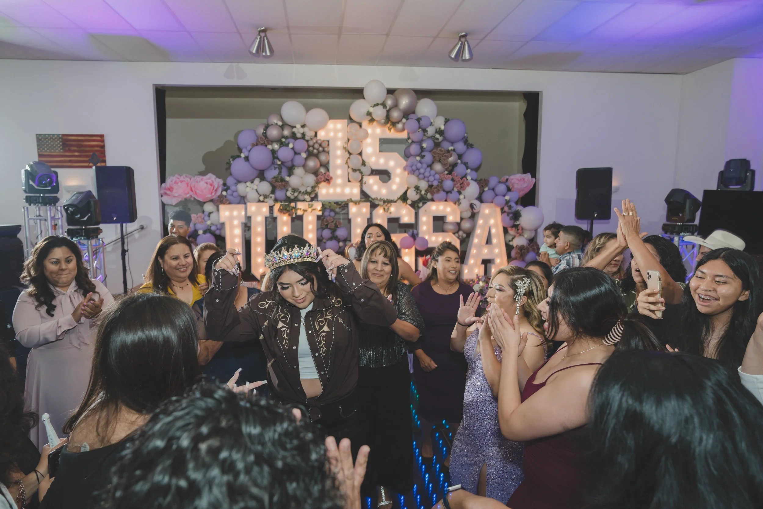 A group of women and children celebrating at a party with a large decorative backdrop featuring illuminated numbers 15, surrounded by balloons and flowers.