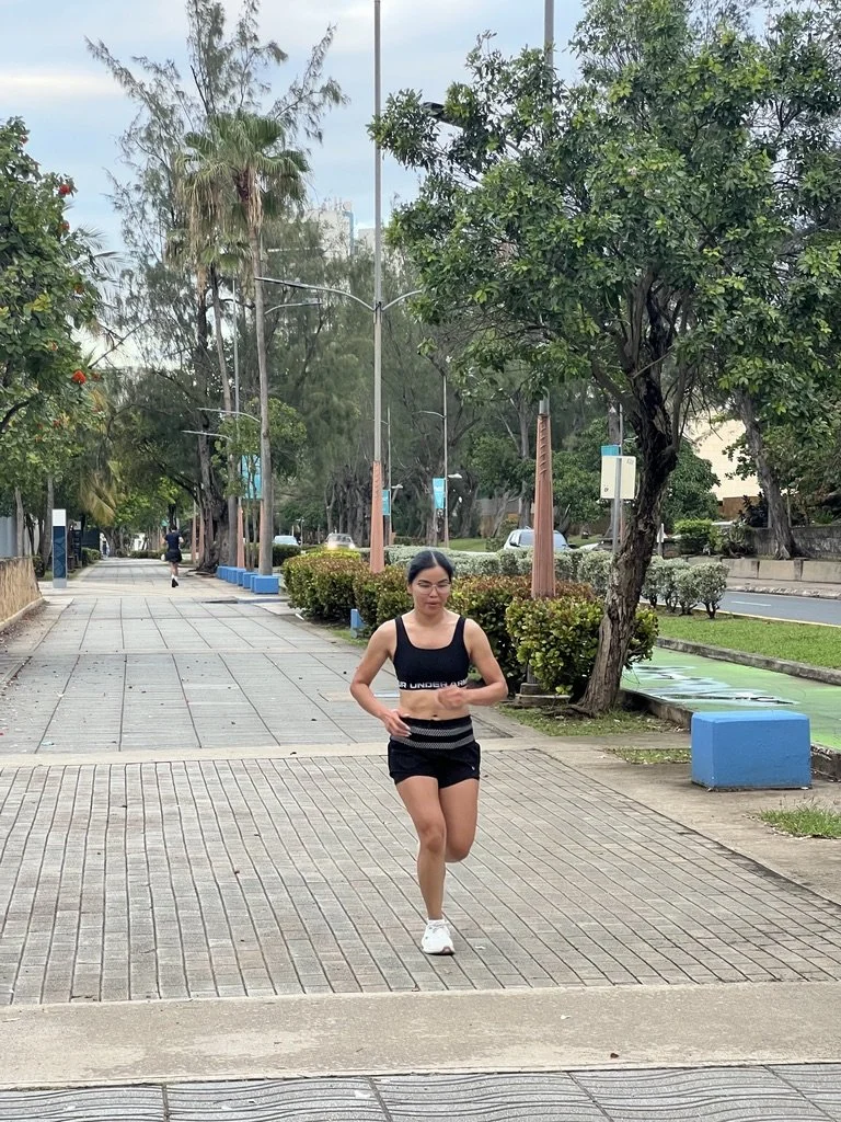 A woman jogging on a sidewalk in a park-like area with trees and shrubs, wearing a black sports bra, black shorts, and white sneakers.
