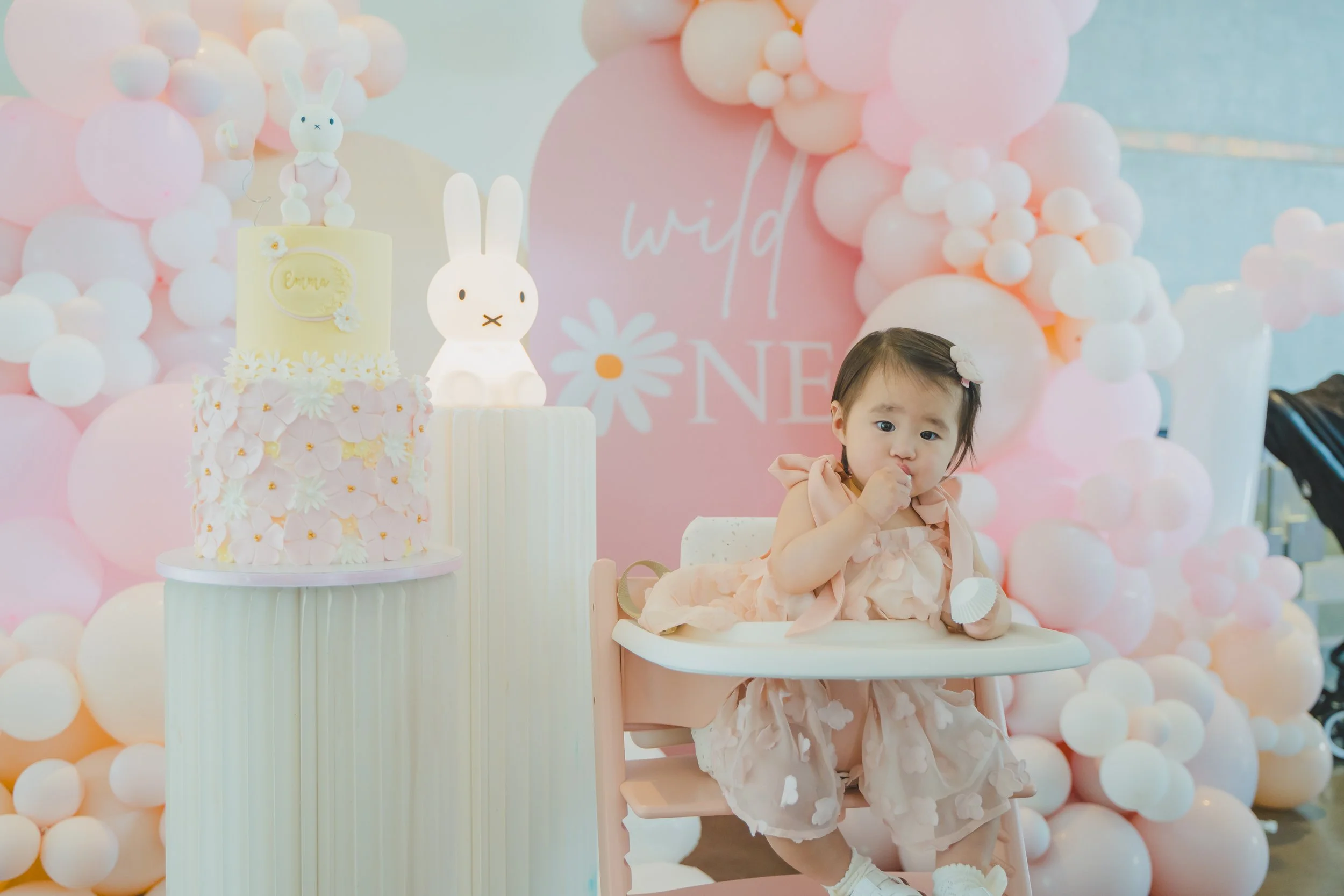 A young girl in a peach-colored dress with tulle skirt sits in a high chair at a birthday celebration. The background features pink and white balloon arrangements, a pink daisy-themed backdrop with the words 'wild ONE,' a whimsical white rabbit lamp,