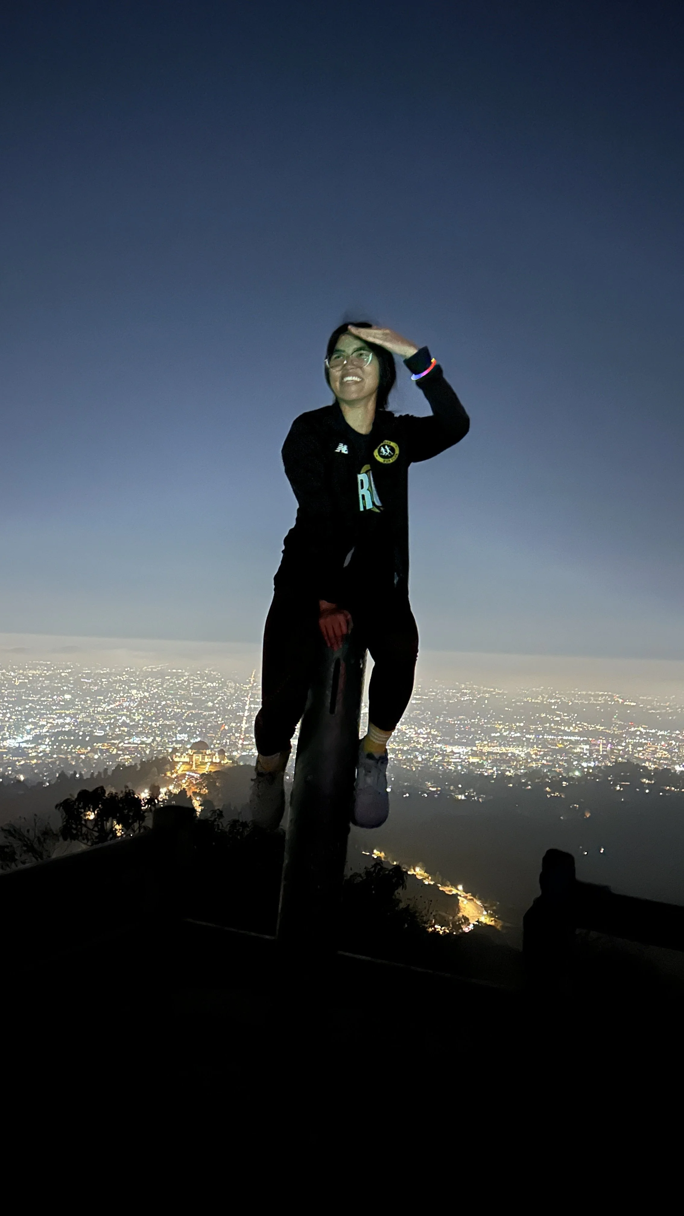 Person standing on a mountain peak during dusk, holding a hand to their forehead, with a cityscape below illuminated with lights.