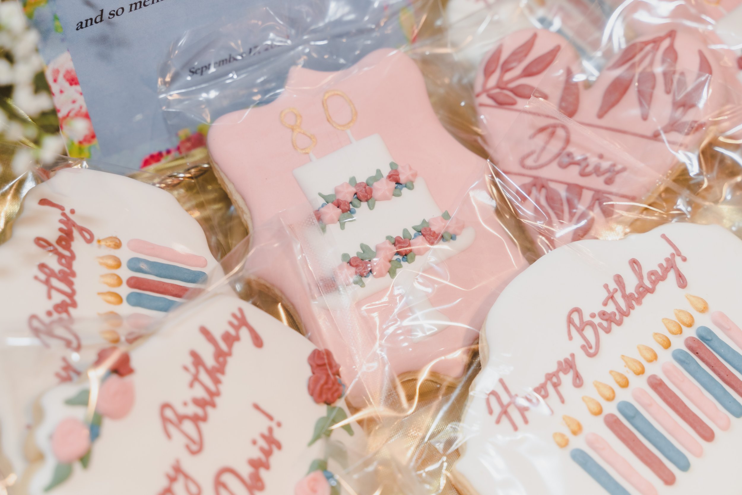 Decorated cookies with birthday themes, including a pink cookie with a cake and candles, celebrating a 80th birthday, wrapped in clear plastic, with some cookies featuring the words 'Happy Birthday'.