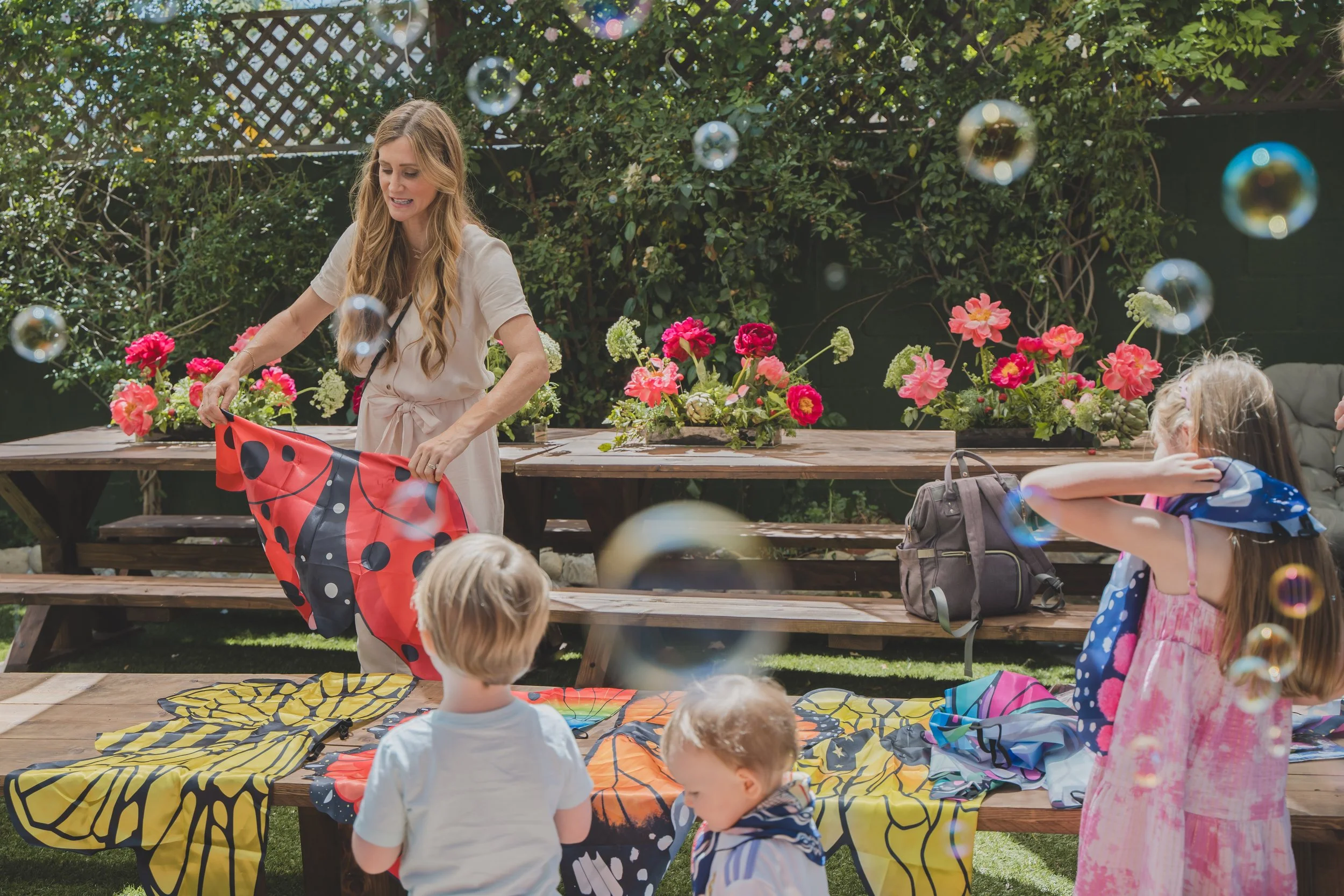 A woman with long blonde hair wearing a beige dress stands in front of children, holding a red and black ladybug kite. The children are sitting on colorful blankets on a wooden table, surrounded by pink flowers and greenery, outdoors. Bubbles float i